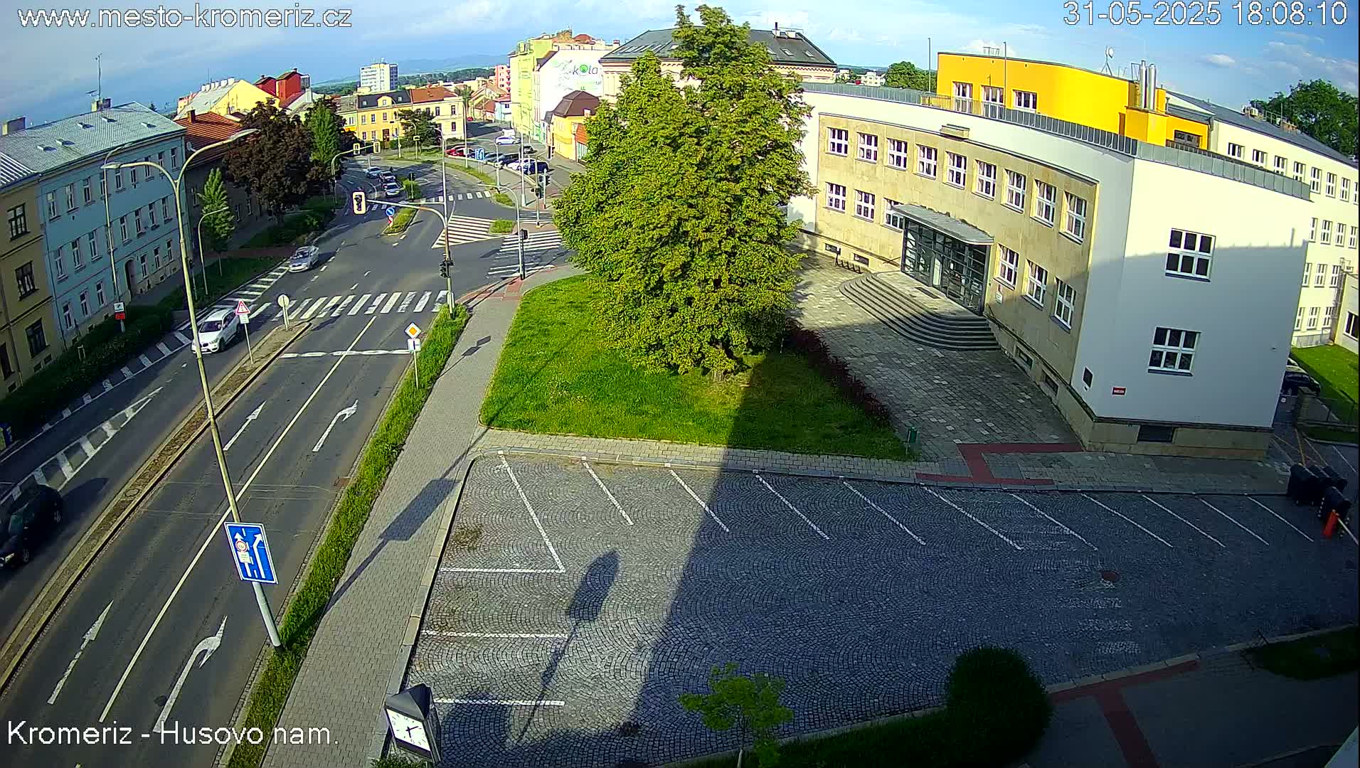 A high-angle, sunny view of a street intersection with buildings and a parking lot, showing cars on the road and a large tree casting a long shadow.