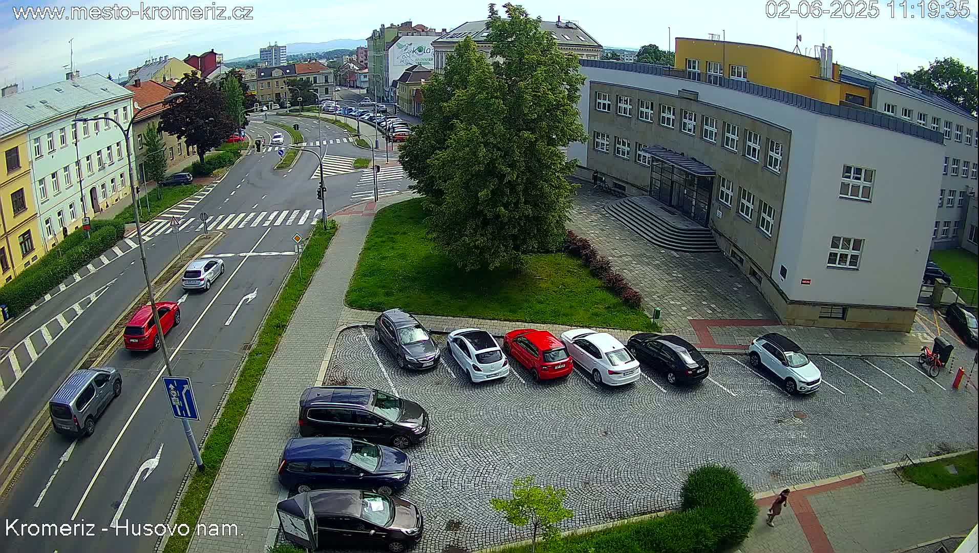 A sunny day shows a street intersection with several cars parked and driving, along with a multi-story building and a large tree in a grassy area.