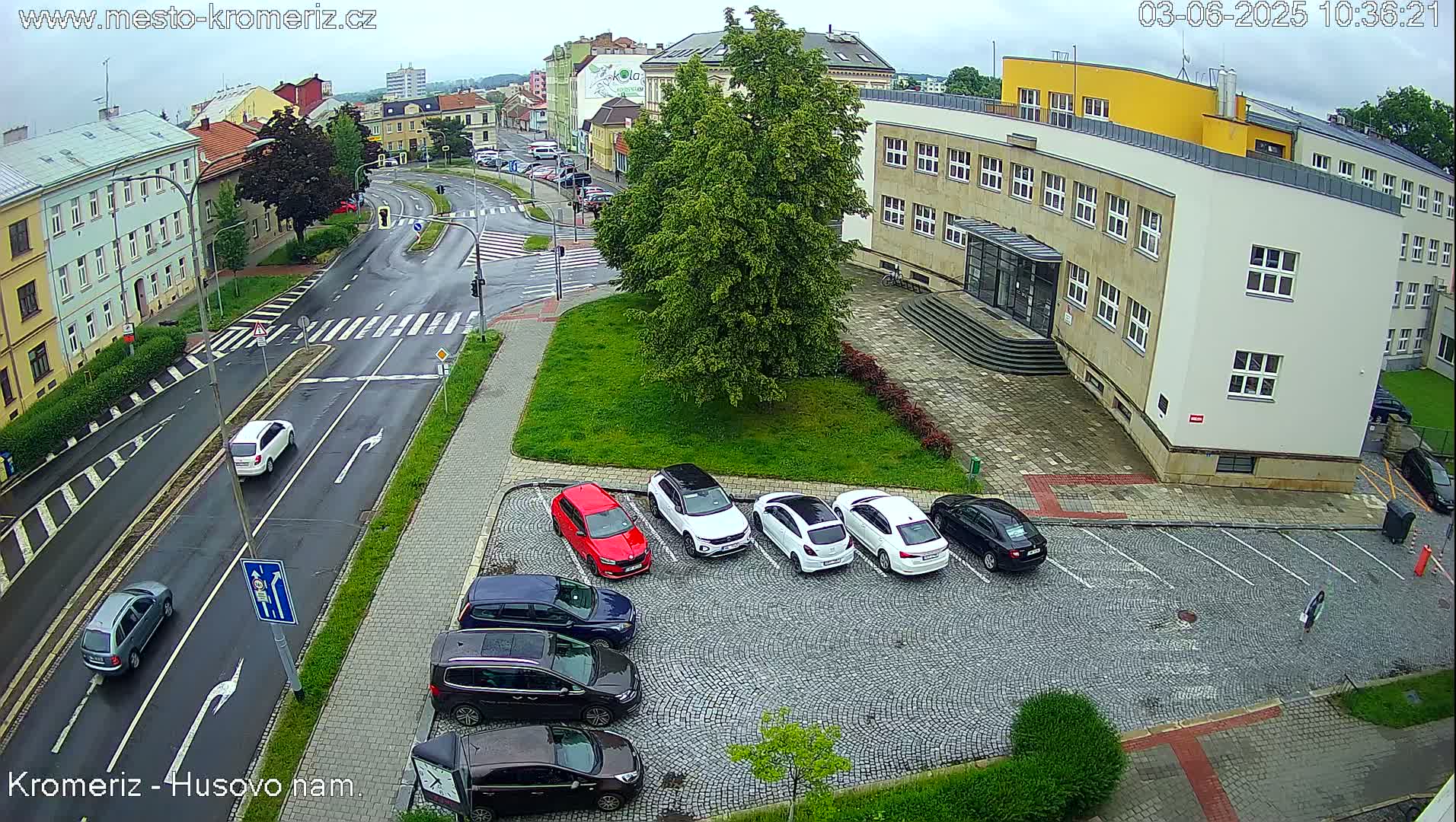 A wet street scene shows several cars parked in a cobblestone lot next to a large building under an overcast sky.
