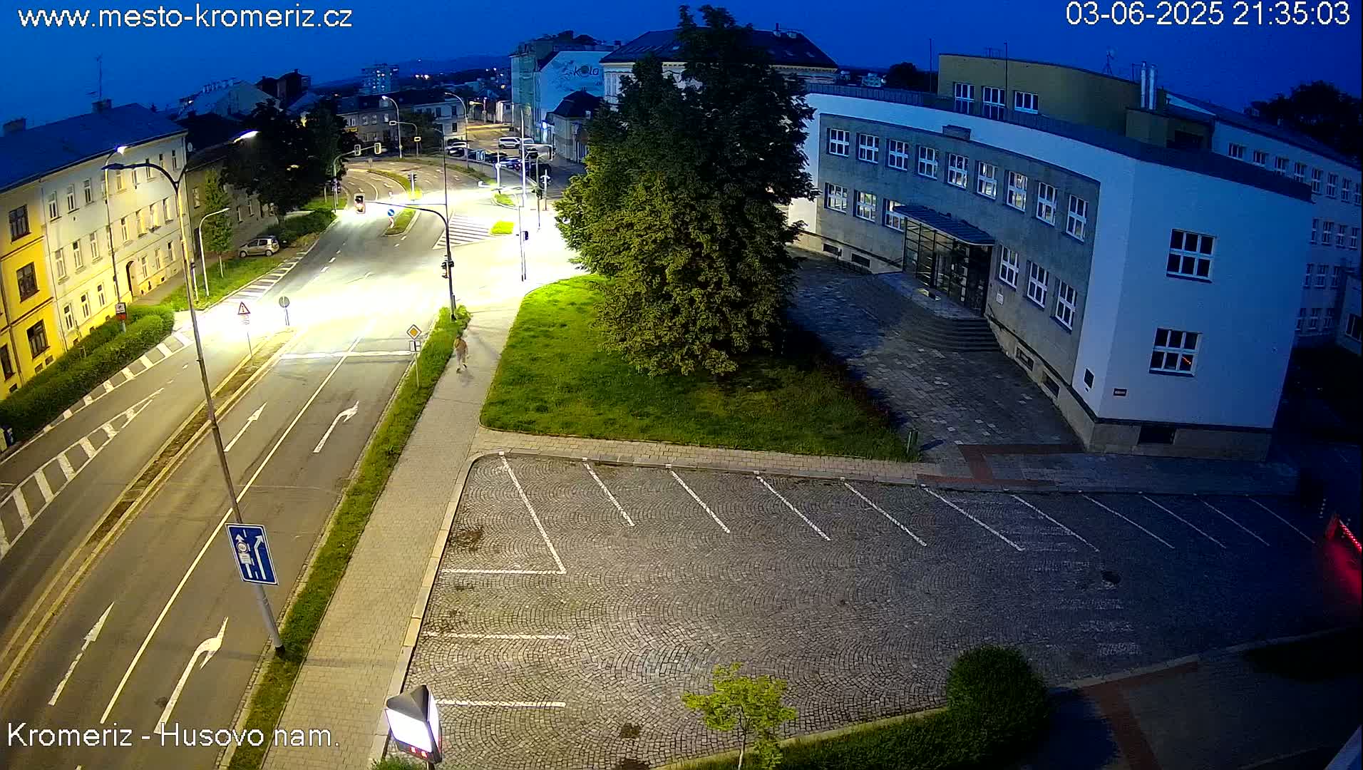 A high-angle nighttime view shows a paved roadway, a parking lot, a large building, and a person walking on a sidewalk under a twilight sky.