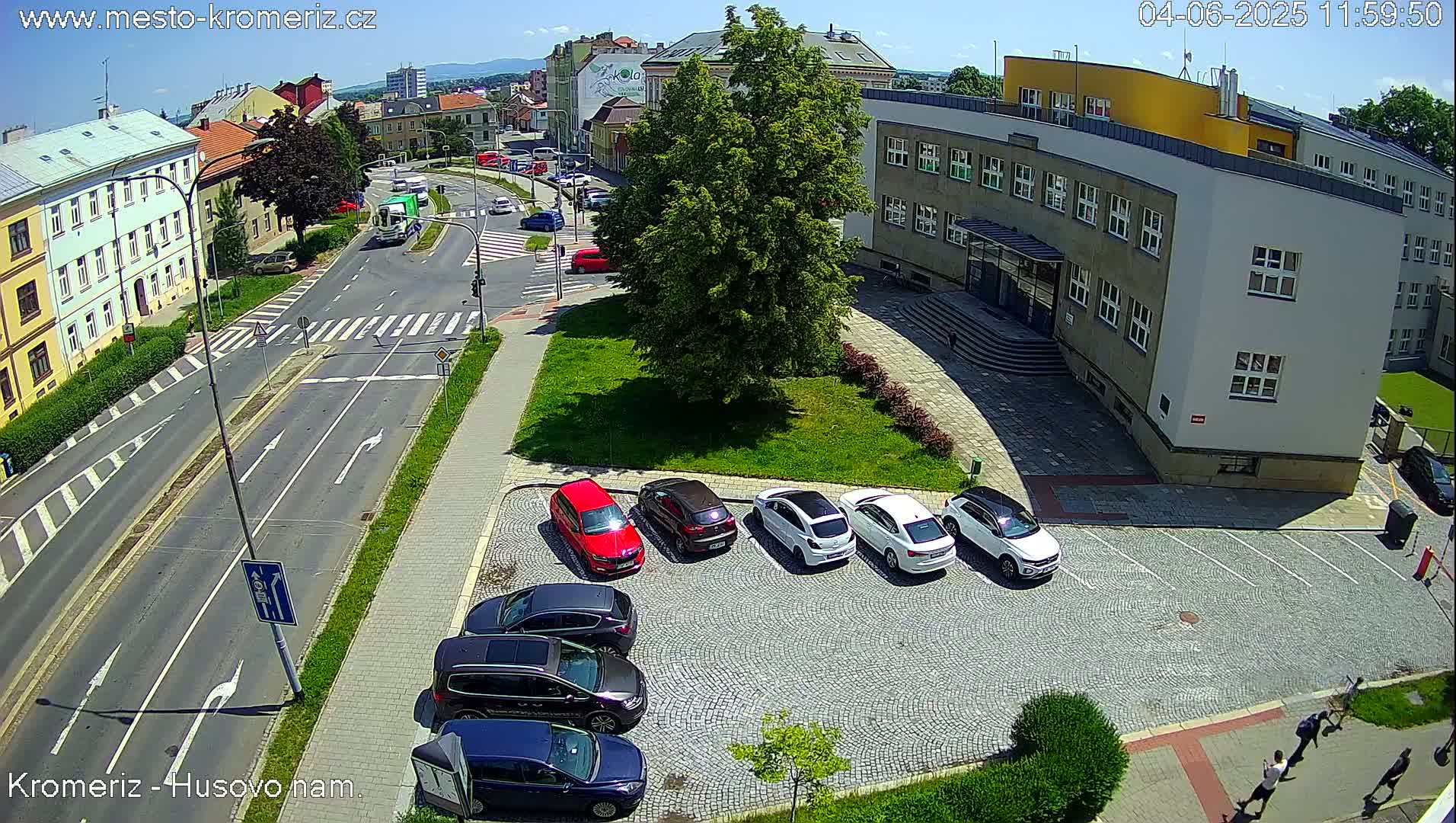 A sunny overhead view shows a paved area with several cars parked near a large building and a street intersection with crosswalks.