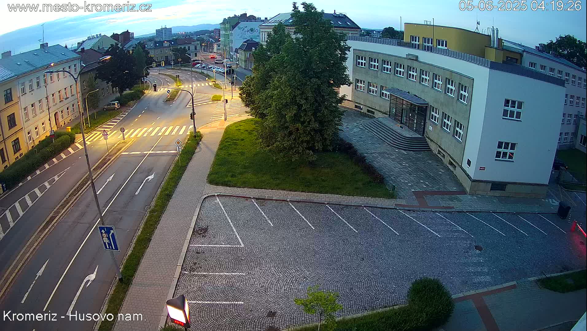 An empty cobblestone parking lot is adjacent to a street intersection at dusk, with a multi-story building in the background.
