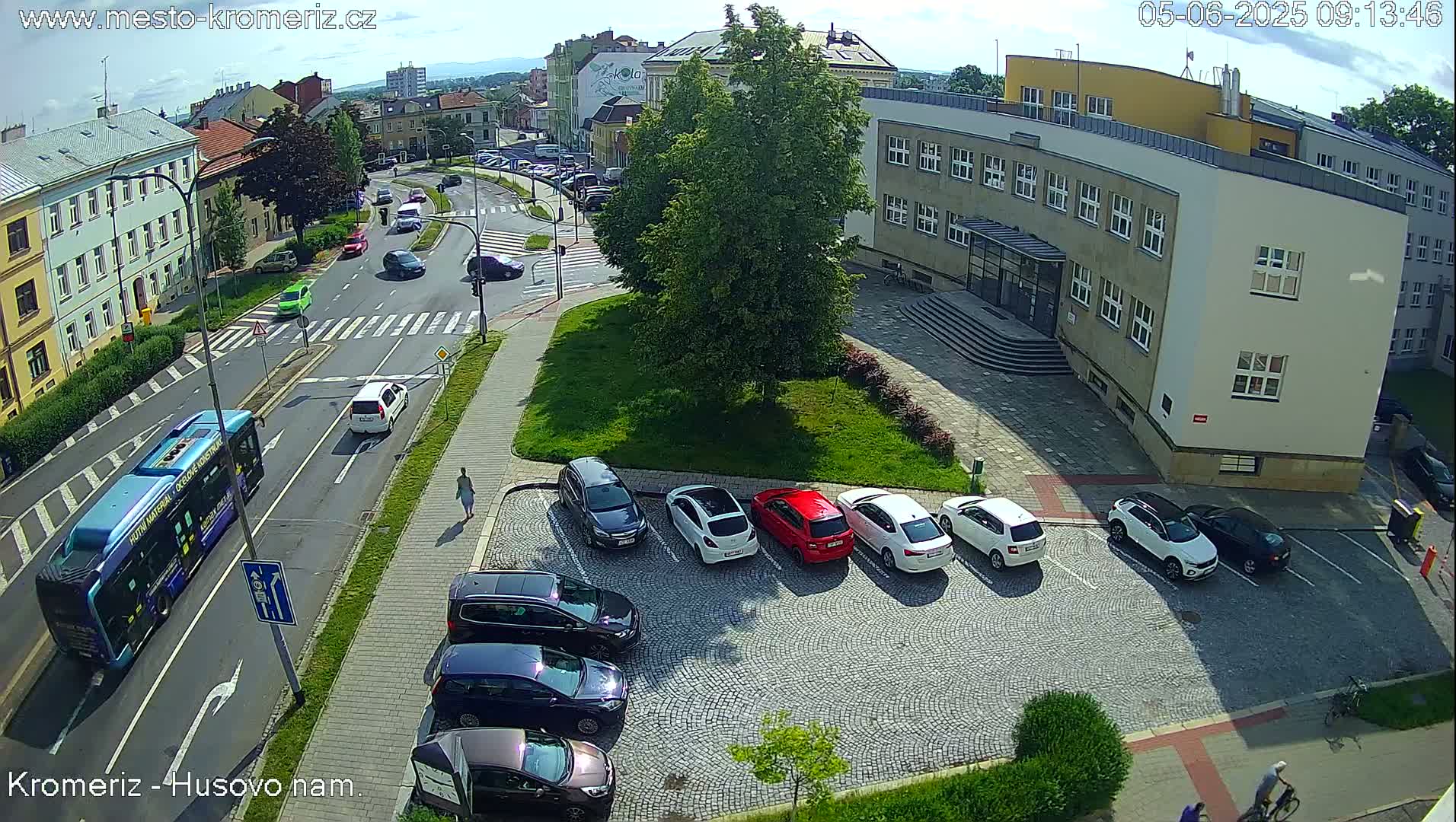 A sunny day shows a high-angle view of a city street with cars parked in a lot next to a building, a bus driving down the road, and several other cars driving.