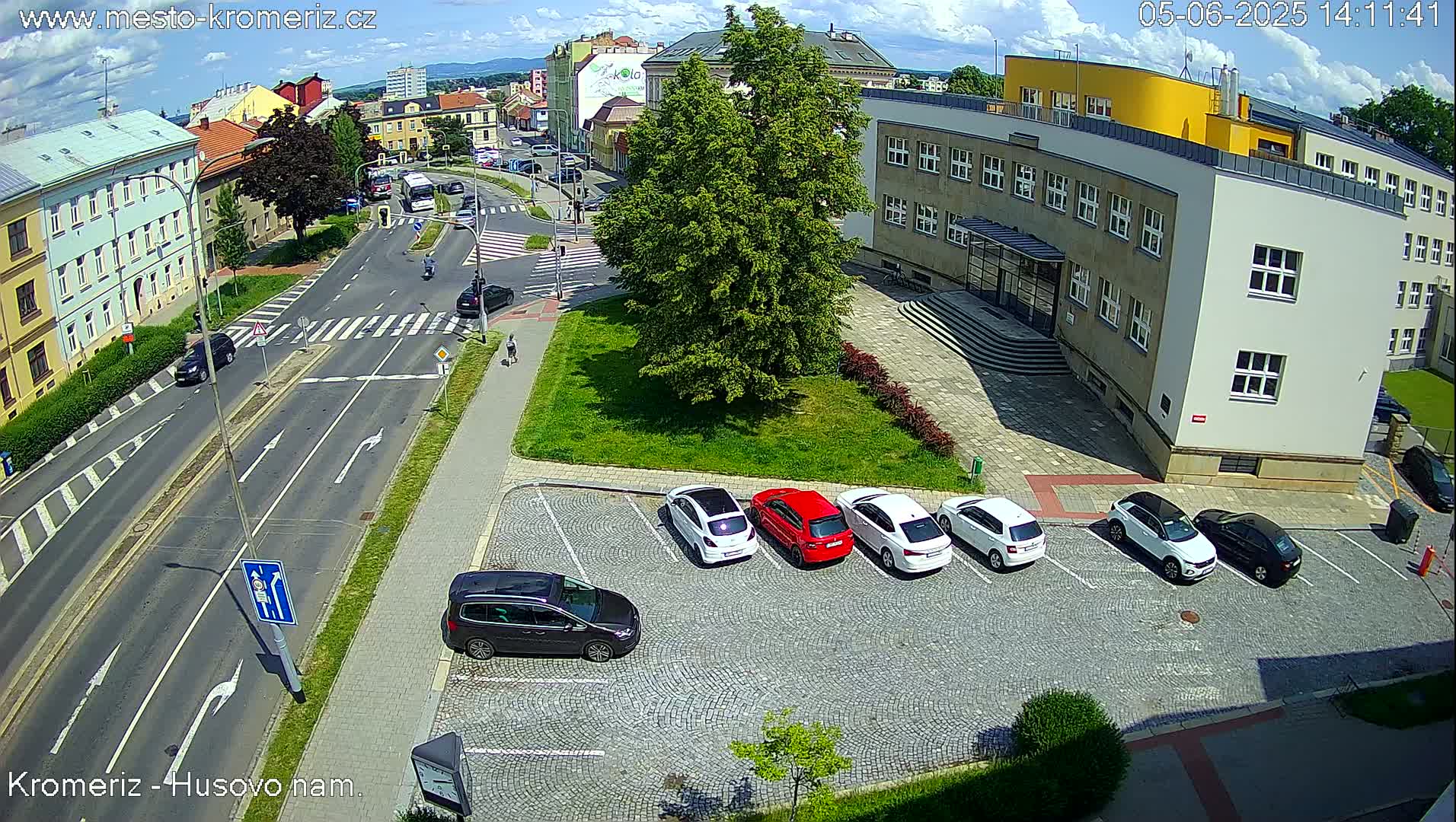 A sunny day reveals a high-angle view of a city intersection with several cars parked in a lot near a large building, and other vehicles traveling on the roads.