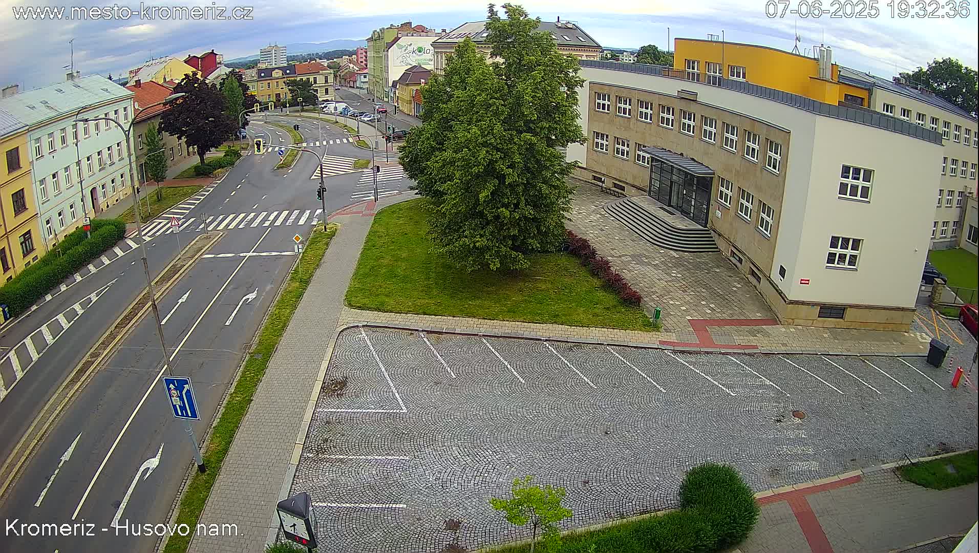 An overhead view shows a paved road intersection with crosswalks, a cobblestone parking lot, and a large building under a partly cloudy sky.