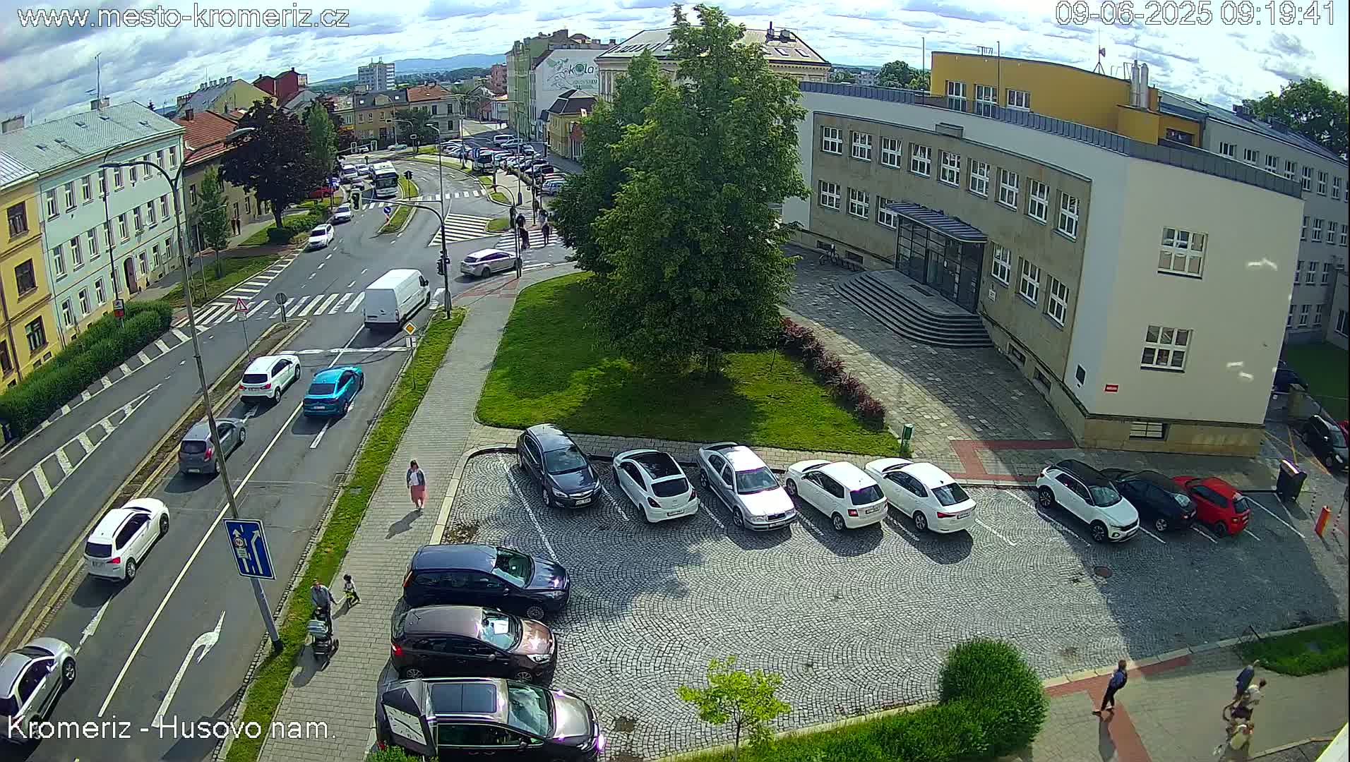 A partly sunny day shows a street intersection with cars parked in a lot next to a large building and several pedestrians walking on sidewalks and streets.