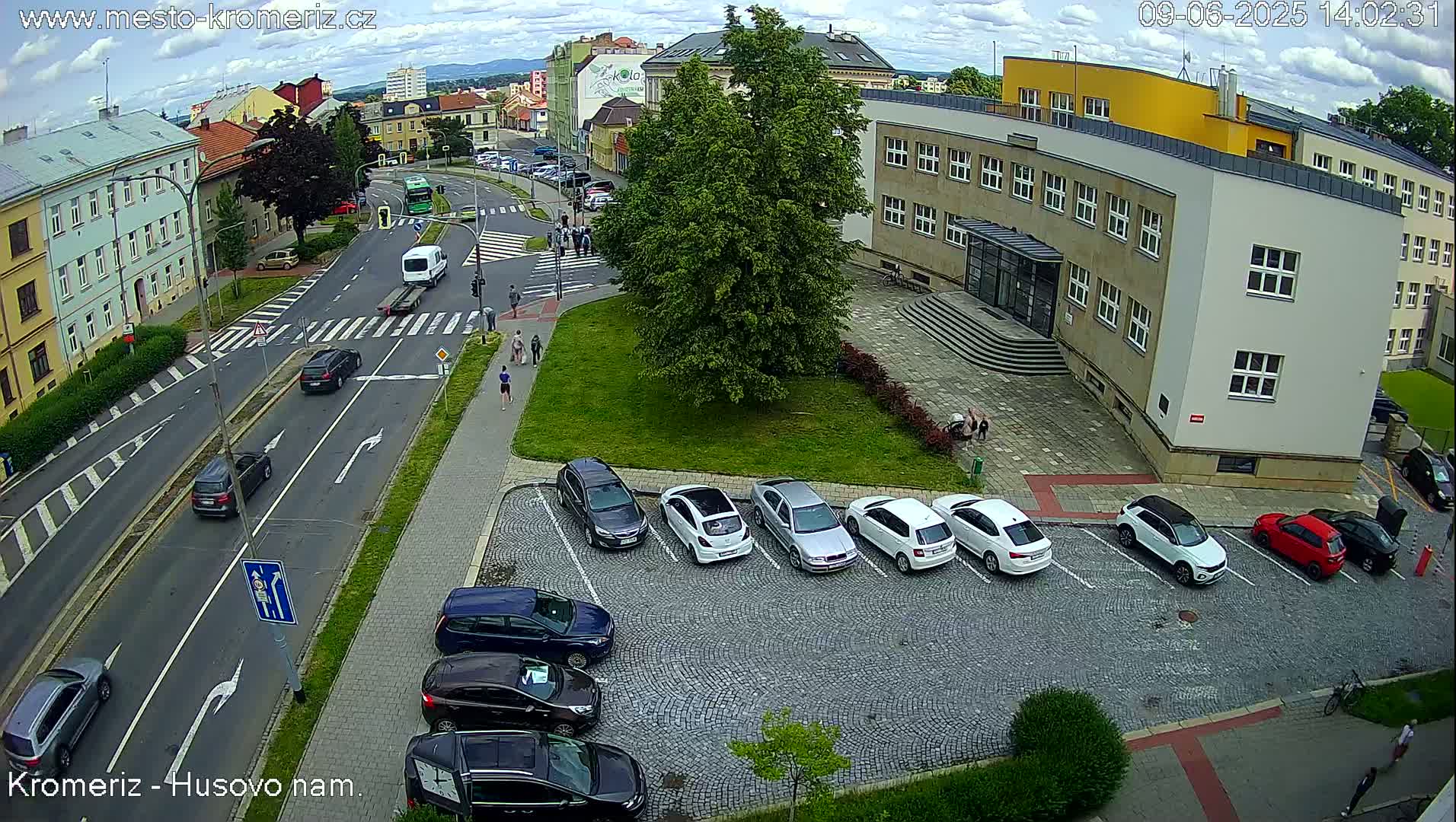A partly cloudy day shows a street scene with cars parked in a lot next to a building and several cars driving on the road.