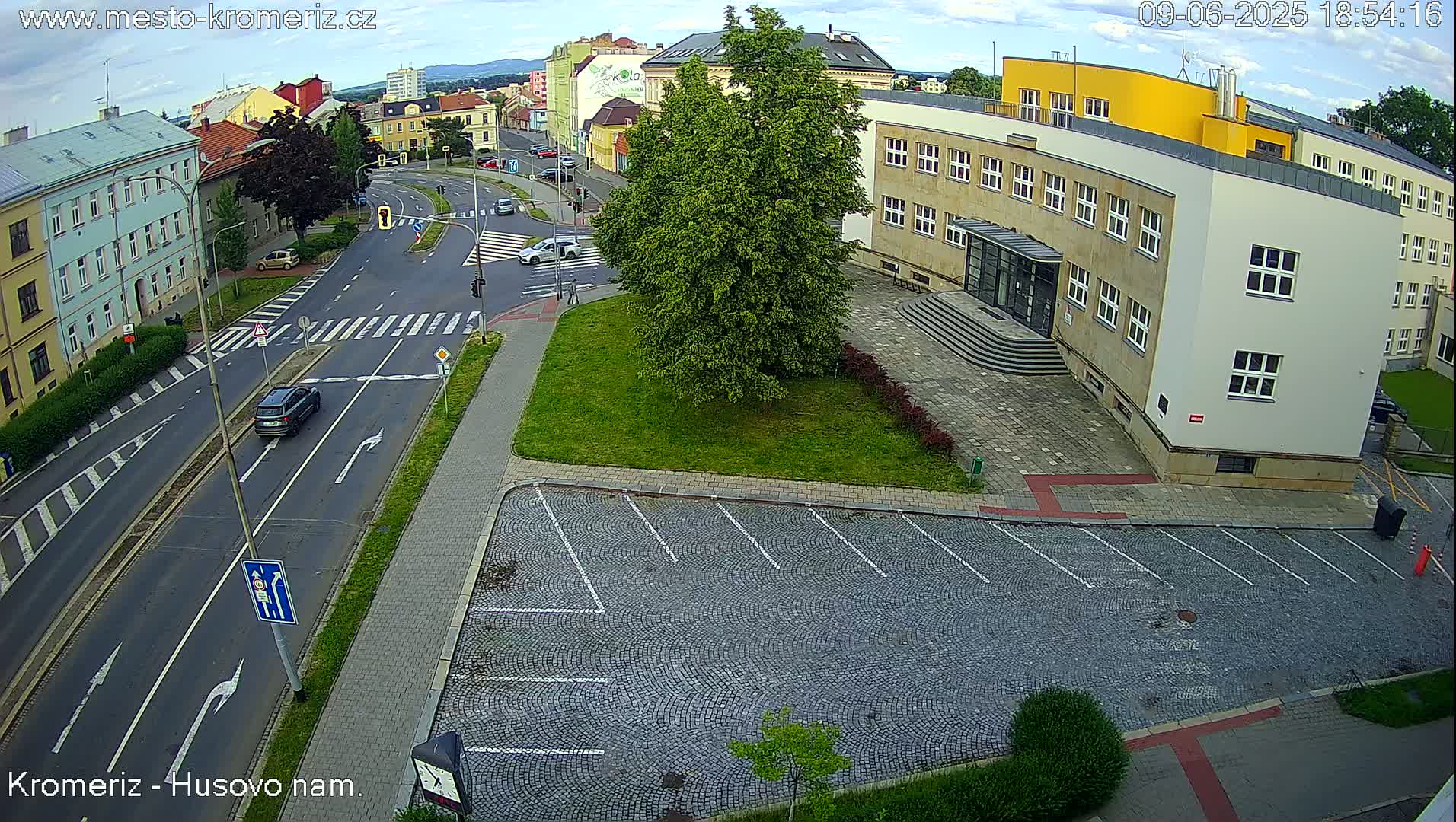 An aerial view shows a street intersection with cars, pedestrian crosswalks, and a large building with a cobblestone parking lot in front, under partly cloudy skies.