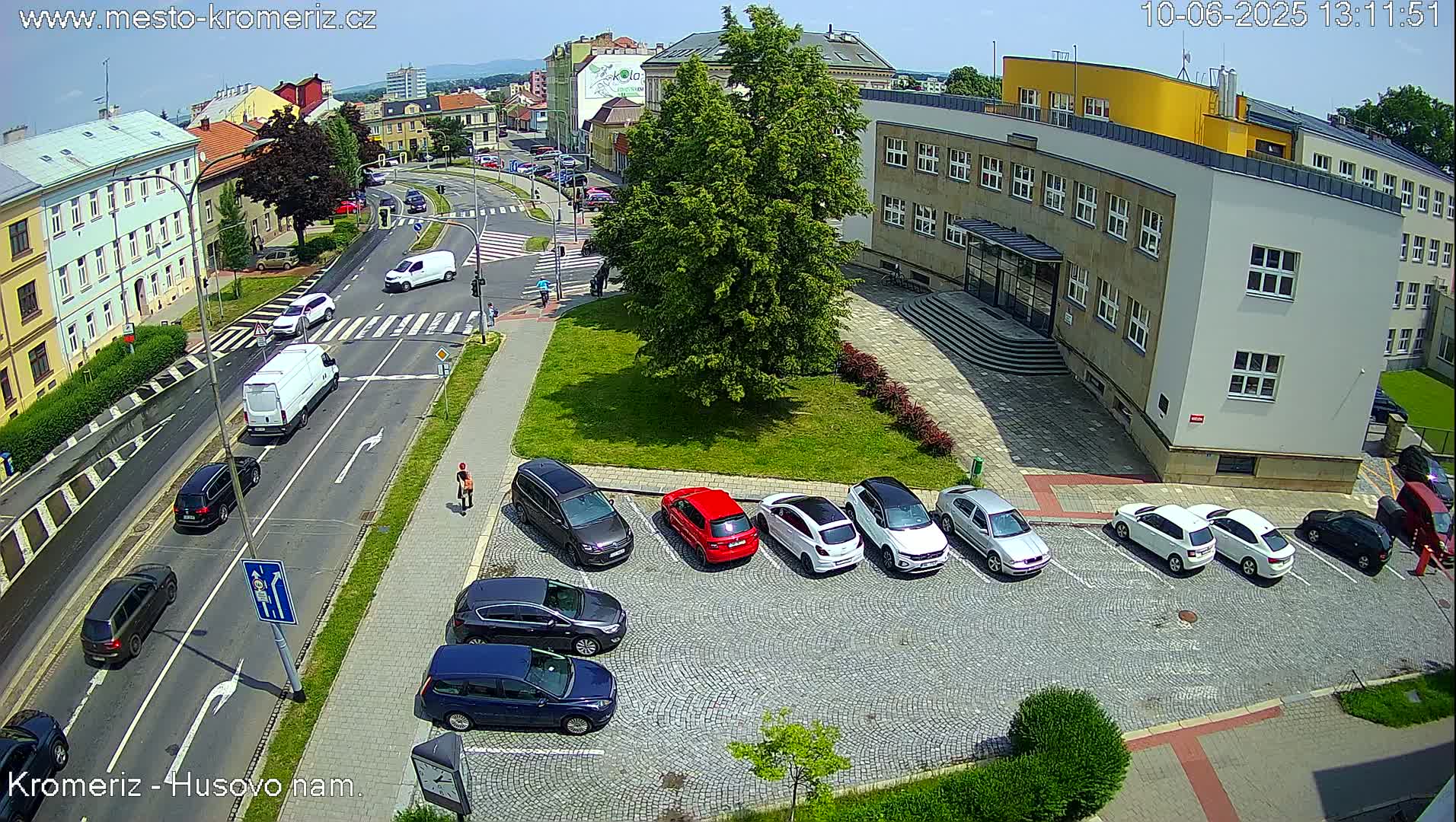 A sunny day shows a city street scene with cars parked in a lot next to a building, and other cars driving on the street.