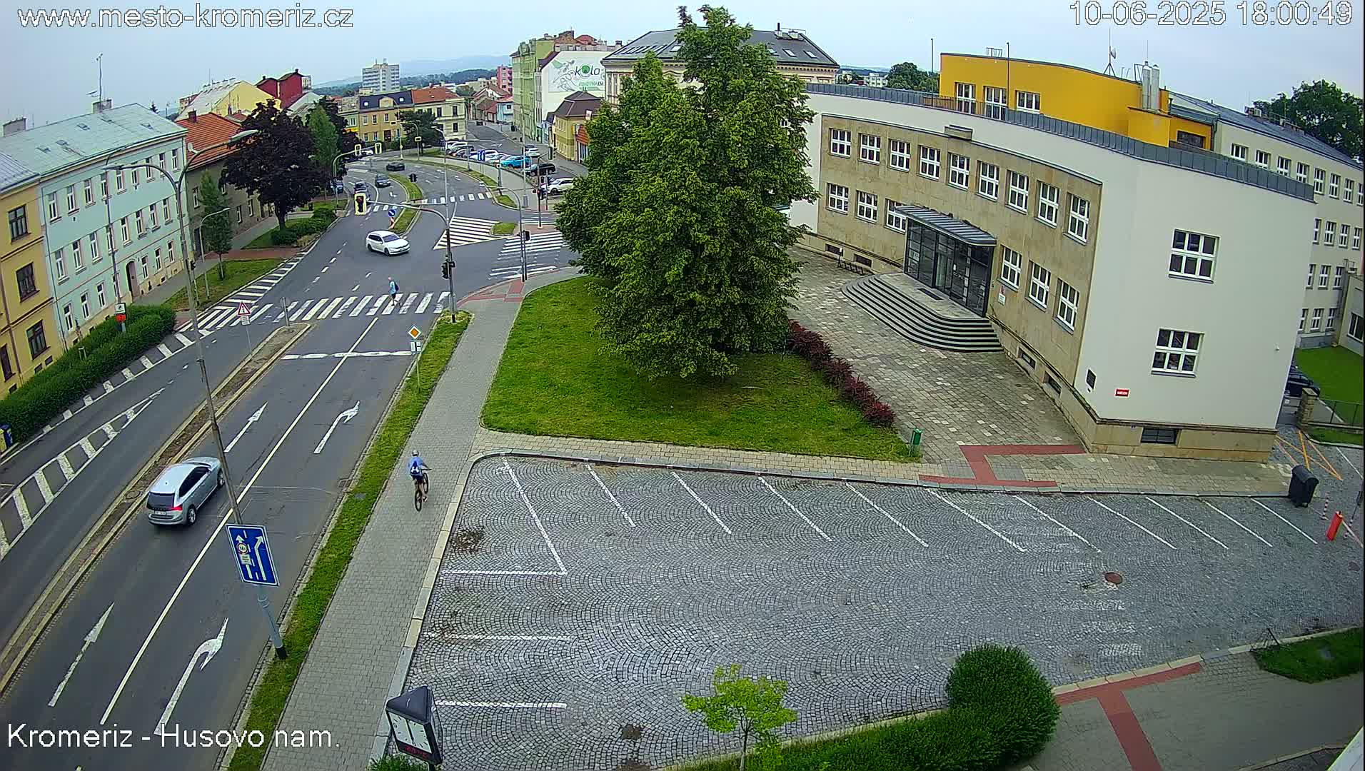 An overcast day reveals a street scene with cars and a bicycle in front of a large building and an empty cobblestone parking lot.