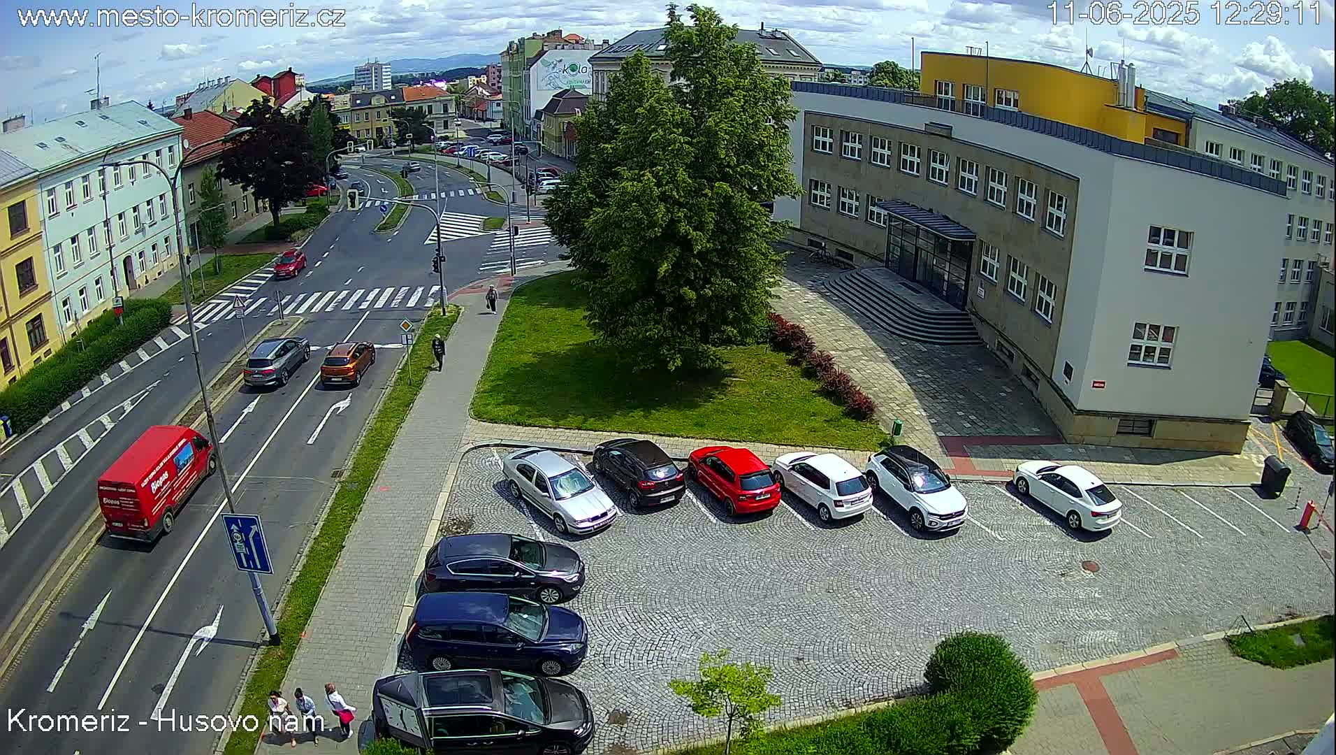 A sunny day reveals a street intersection with several cars driving and parked near a large building and a grassy area.