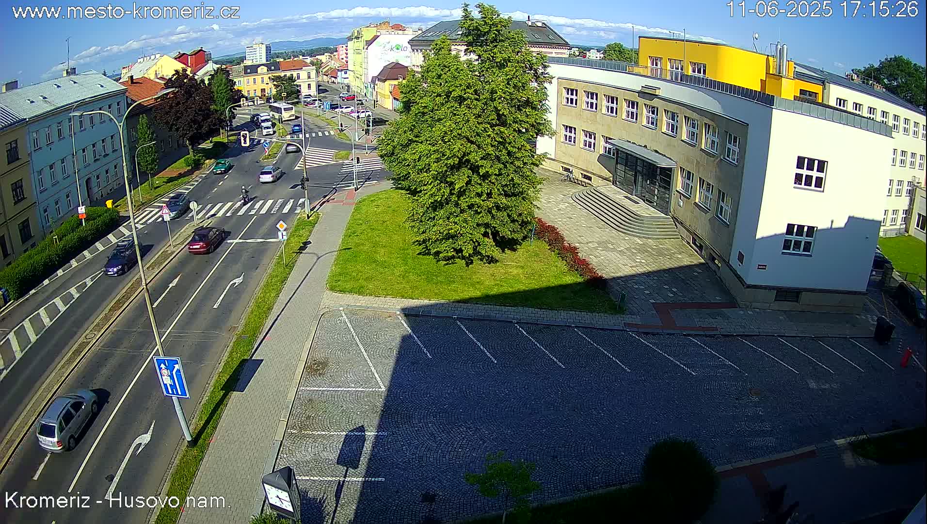 A sunny day reveals a street scene with cars driving on a road next to a large building and a grassy area with a parking lot.