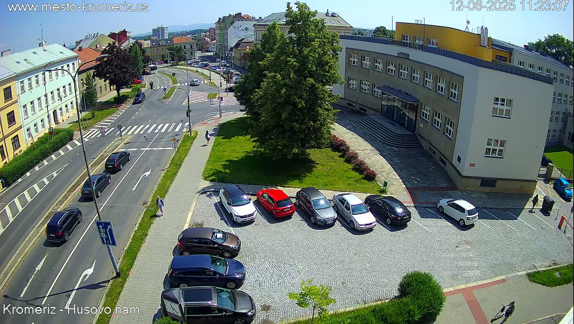 A sunny day view of a city street intersection with several cars parked in a lot next to a building, and other cars driving on the road.
