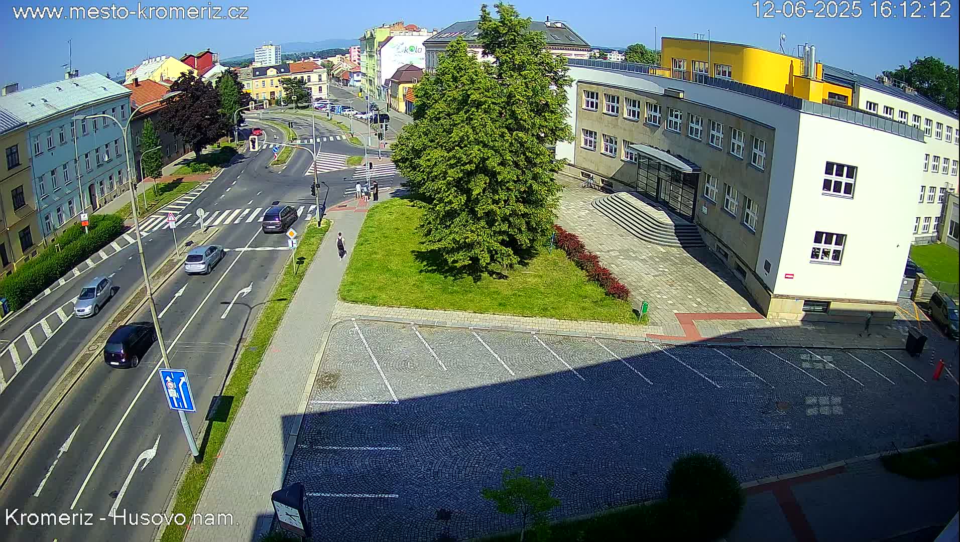 A sunny day view of a street intersection with several cars, pedestrians, and a large building with a parking lot in front.