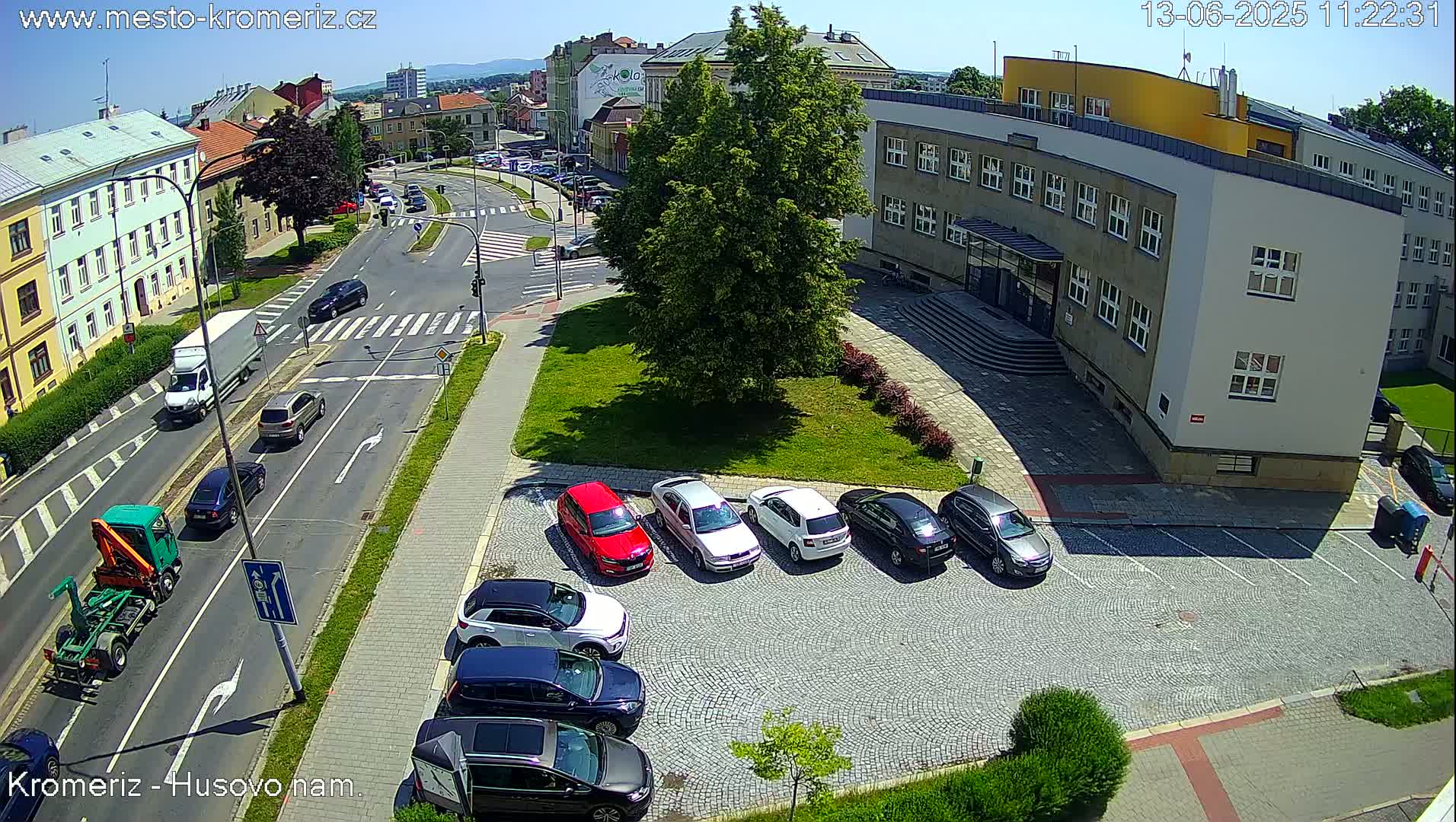 A sunny day overlooking a street intersection with several cars parked in a lot next to a building, and other vehicles traveling down the street.