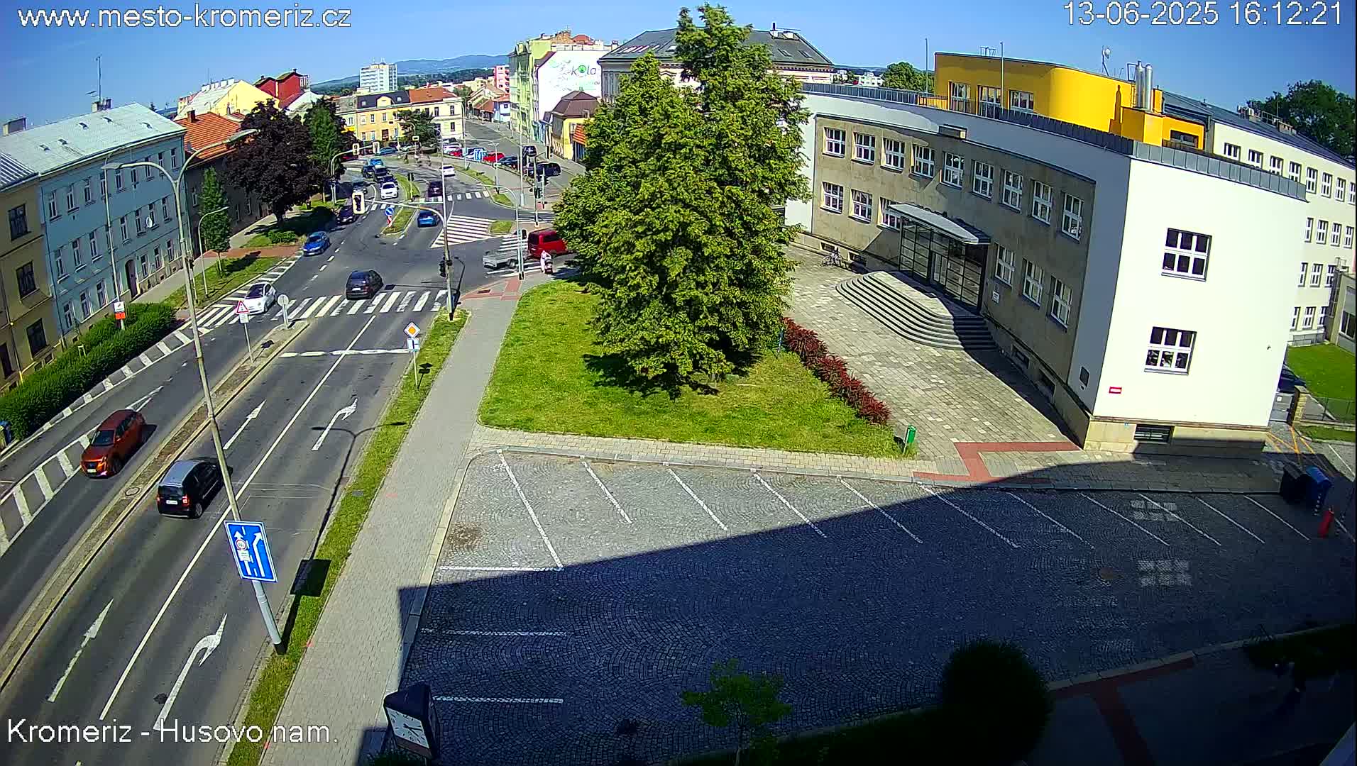 An aerial view of a sunny city street scene shows several cars driving on a multi-lane road that intersects with another road, and a large building with an empty parking lot in front of it.