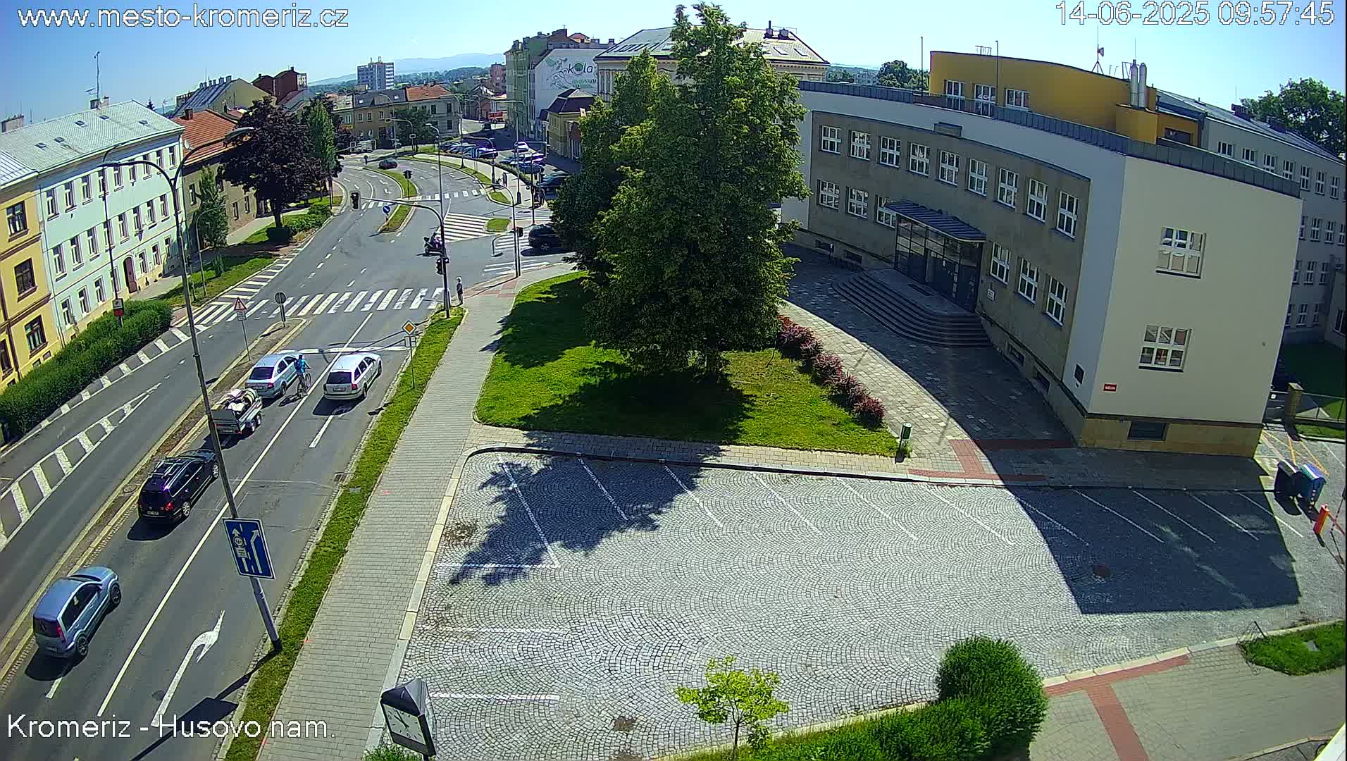 A high-angle, sunny view of a city intersection with several cars, a pedestrian, and a cyclist, next to a large building with a cobblestone parking lot and a grassy area.