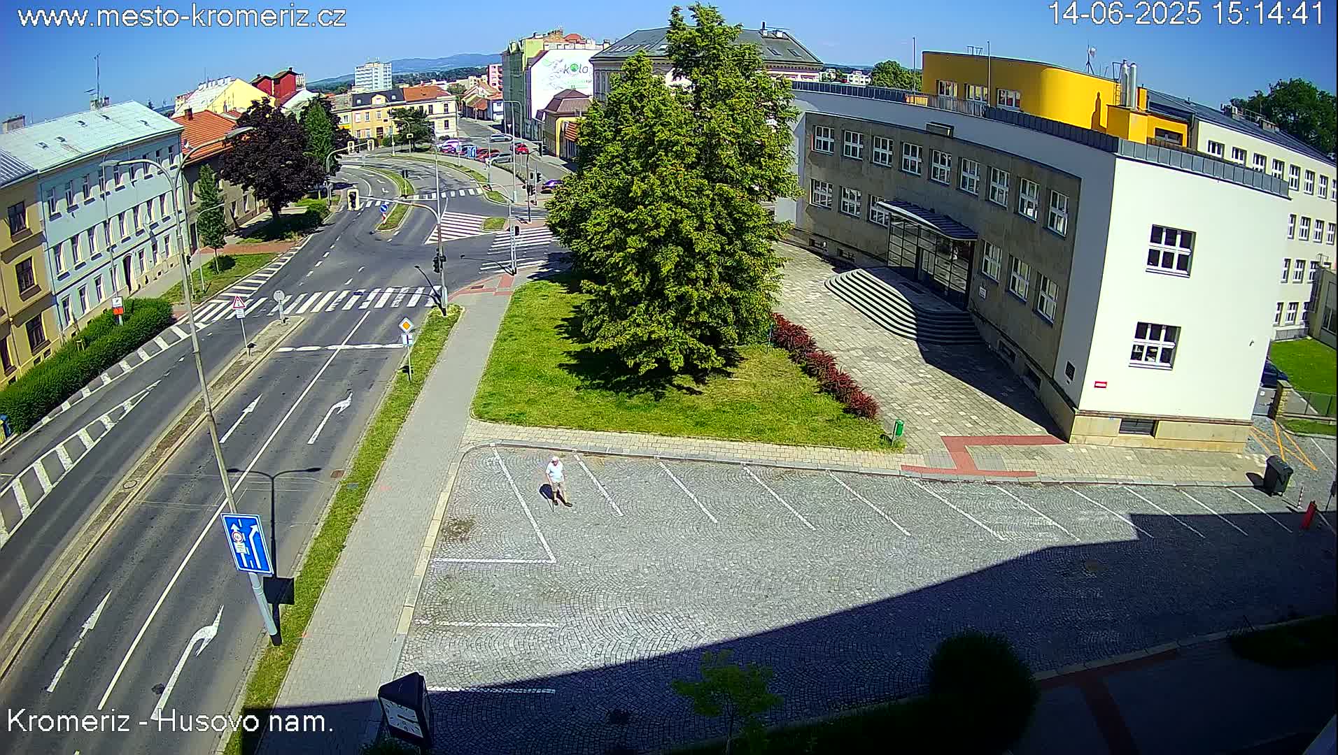 A sunny aerial view shows a street intersection with a large building, a small grassy area with a tree, and a person walking in a paved parking lot.