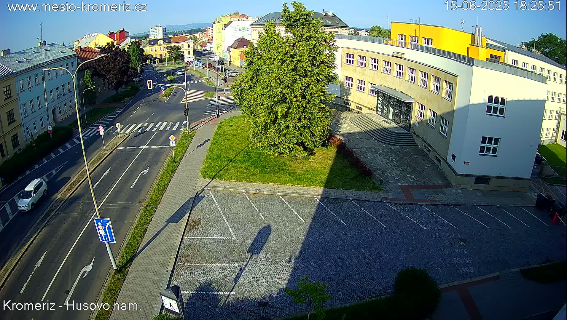 A sunny aerial view shows a street intersection with a large building and an empty cobblestone parking lot.