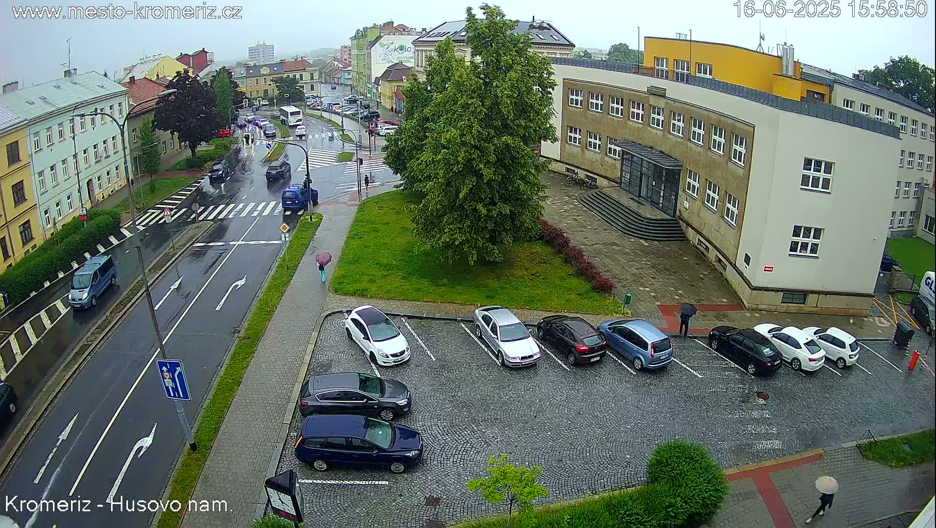A rain-soaked street scene shows several cars parked in a lot next to a multi-story building, with additional cars driving on the wet road.