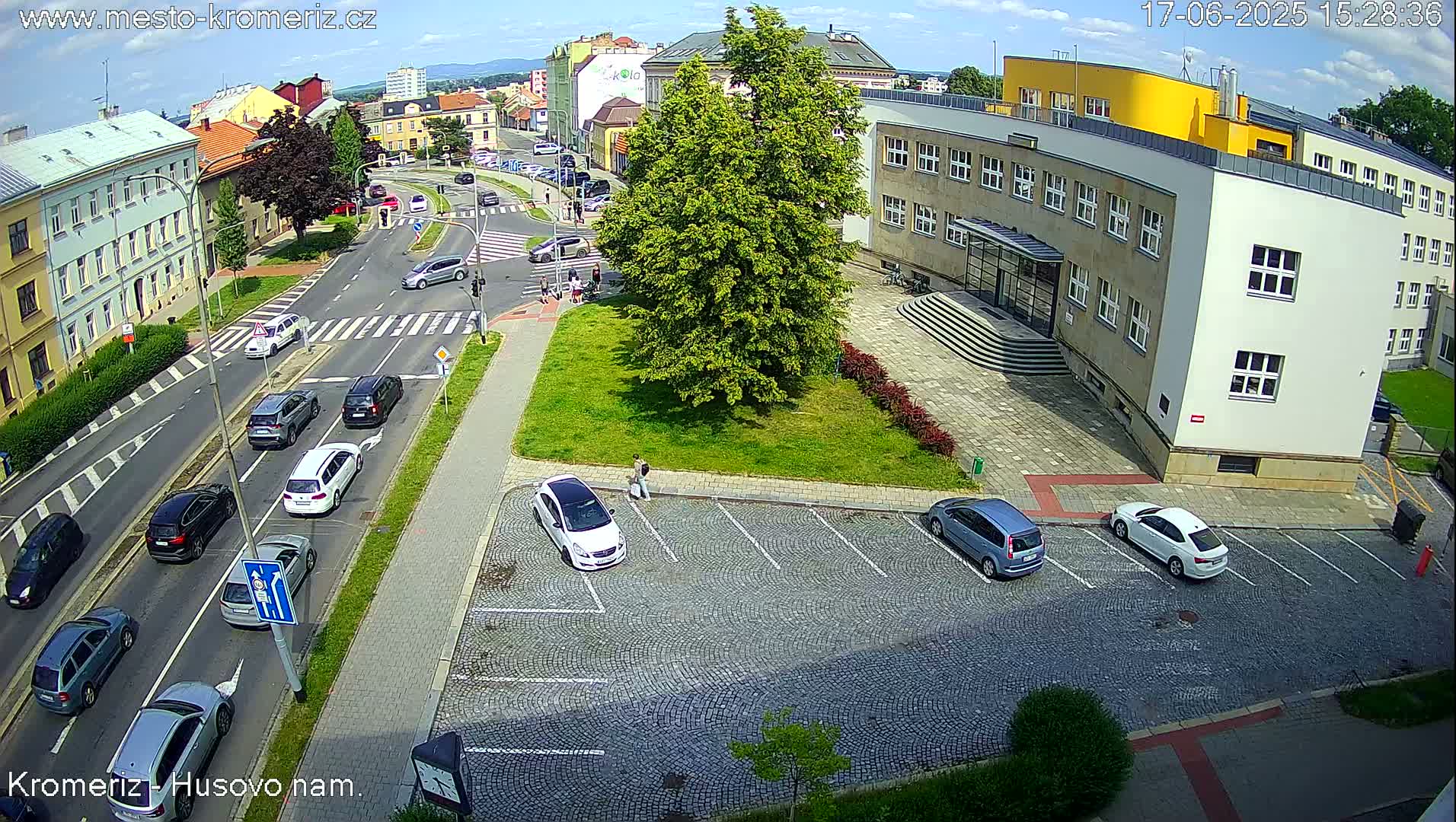 An aerial view shows a sunny day with several cars parked in a cobblestone lot near a multi-story building and a street intersection with multiple cars.