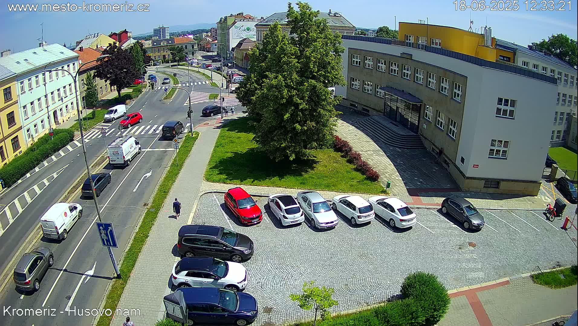 A sunny day view of a city street intersection with cars parked in a lot next to a large building, and several vehicles driving on the roads.