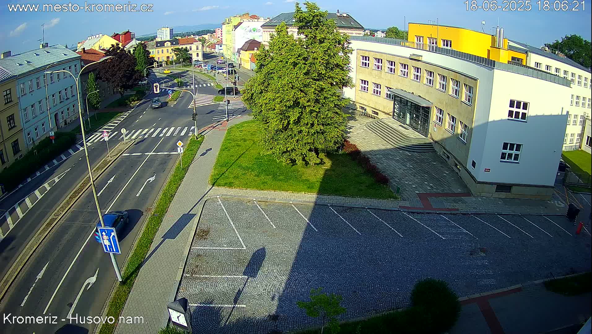 A sunny overhead view of a city street intersection with a large building, a grassy area, and a cobblestone parking lot.