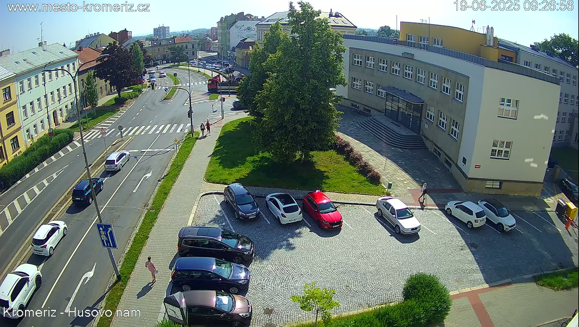 A sunny day shows a street scene with several cars parked in a cobblestone lot next to a building, and other cars driving on a road with pedestrian crossings.