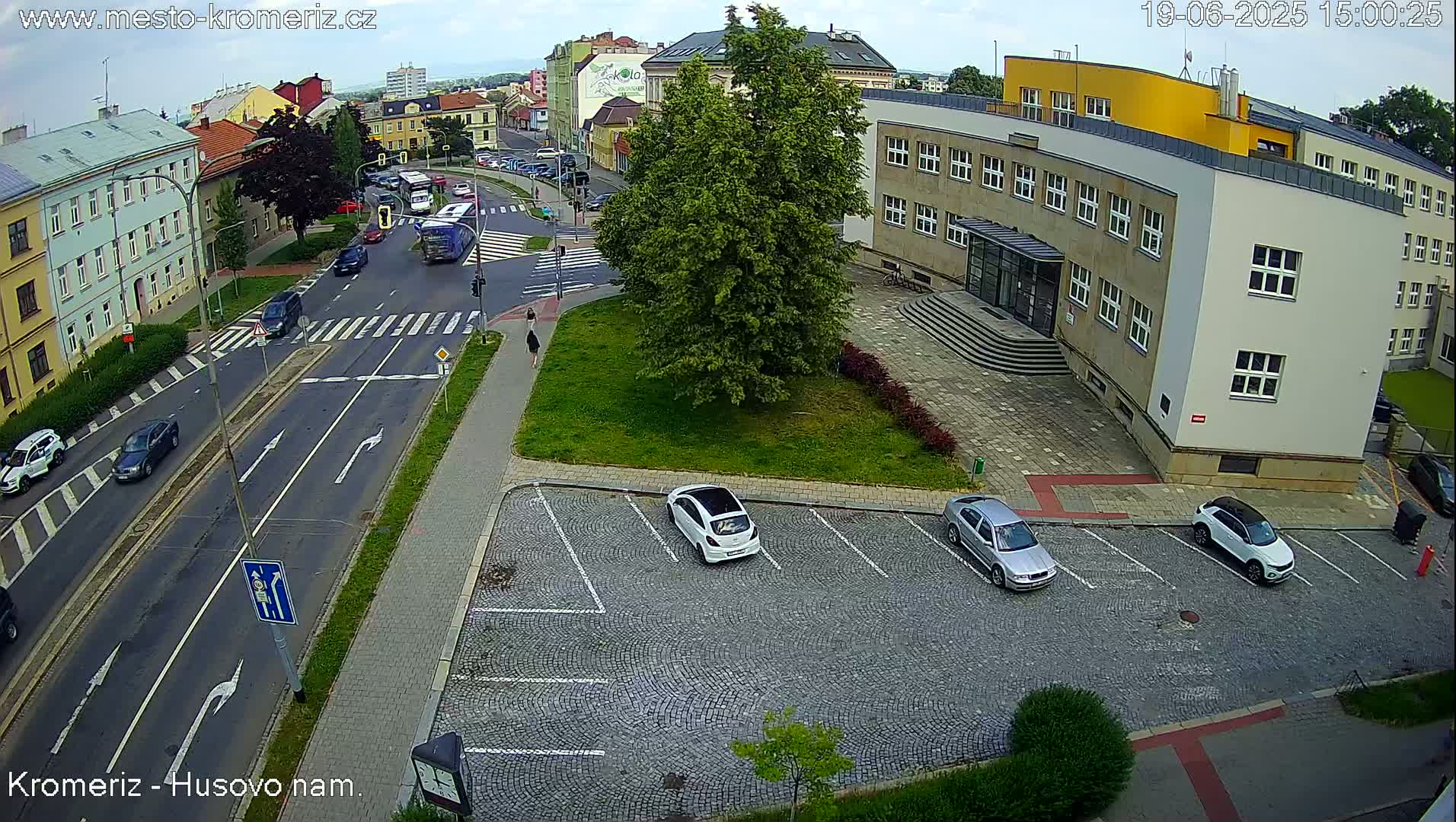 An overcast day shows a street intersection with several cars parked in a cobblestone lot next to a large building, and other cars driving on the streets.
