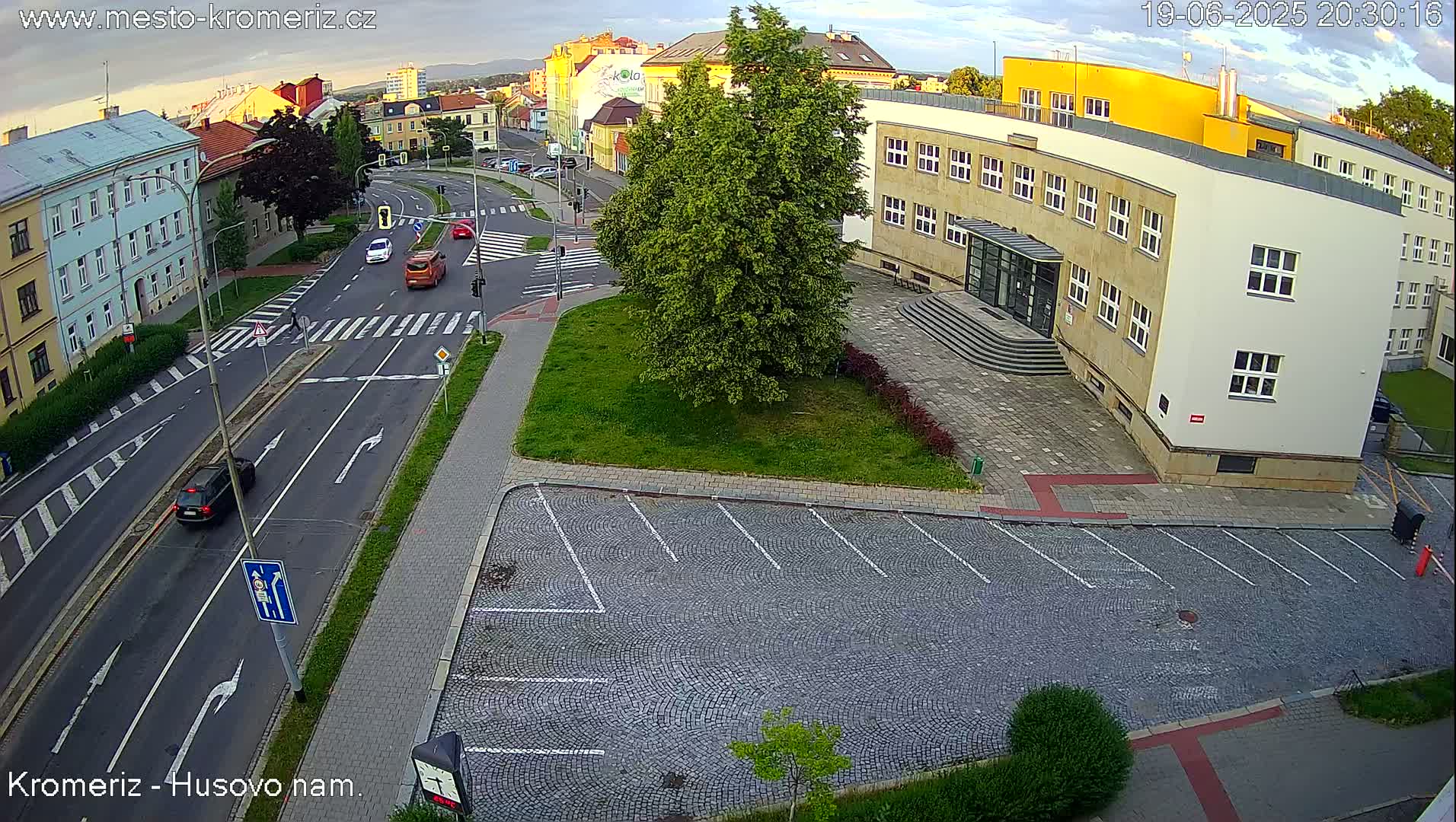An empty cobblestone parking lot is adjacent to a street intersection with a few cars and buildings under a partly cloudy evening sky.