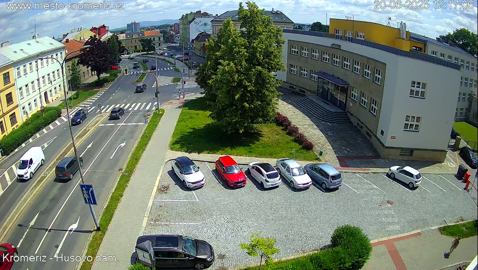 A sunny day shows a street intersection with cars driving on the road and several cars parked in a cobblestone lot next to a large building.