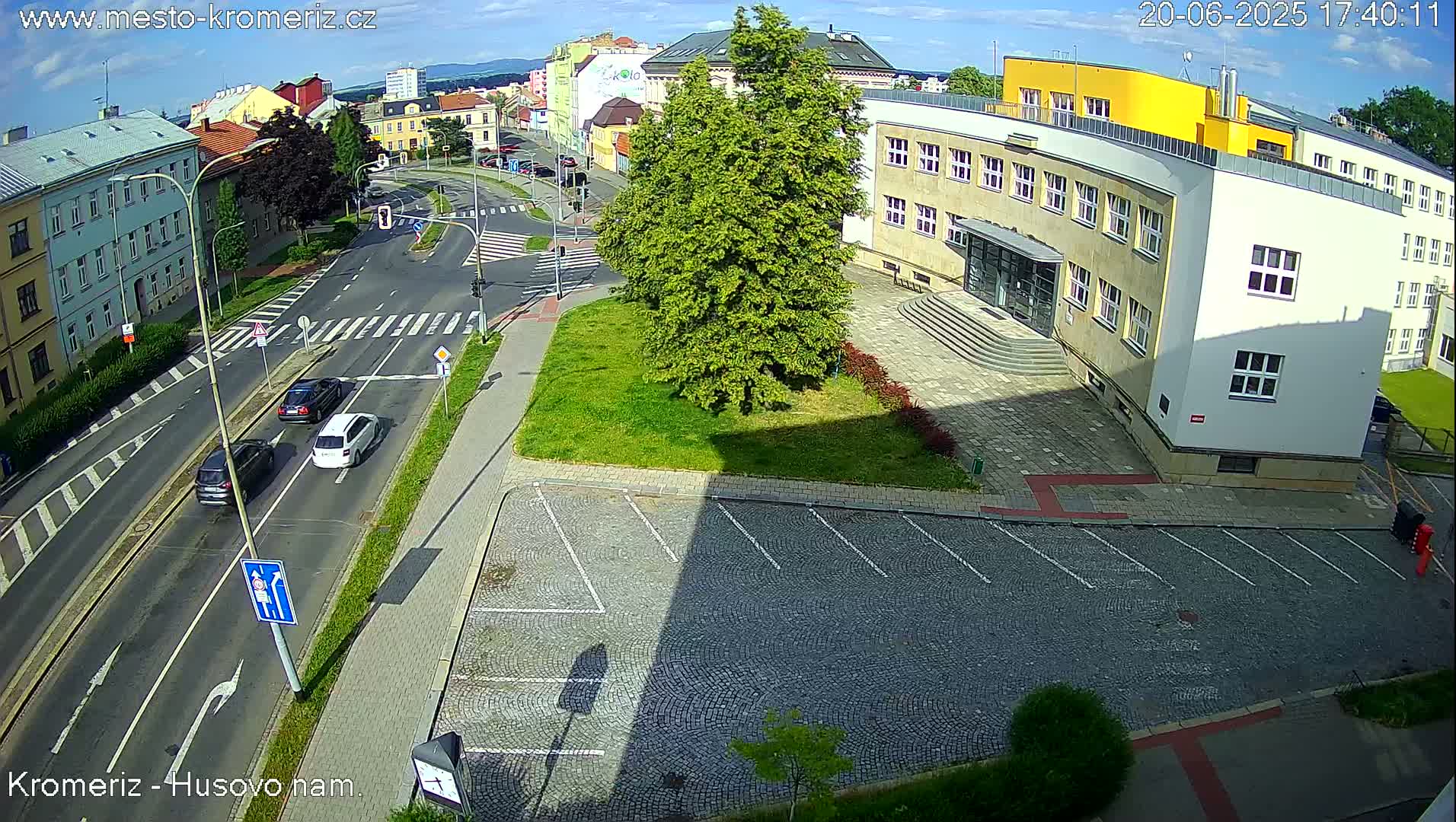 A sunny overhead view shows a street intersection with several cars, a parking lot, and a large building with a grassy area in front.