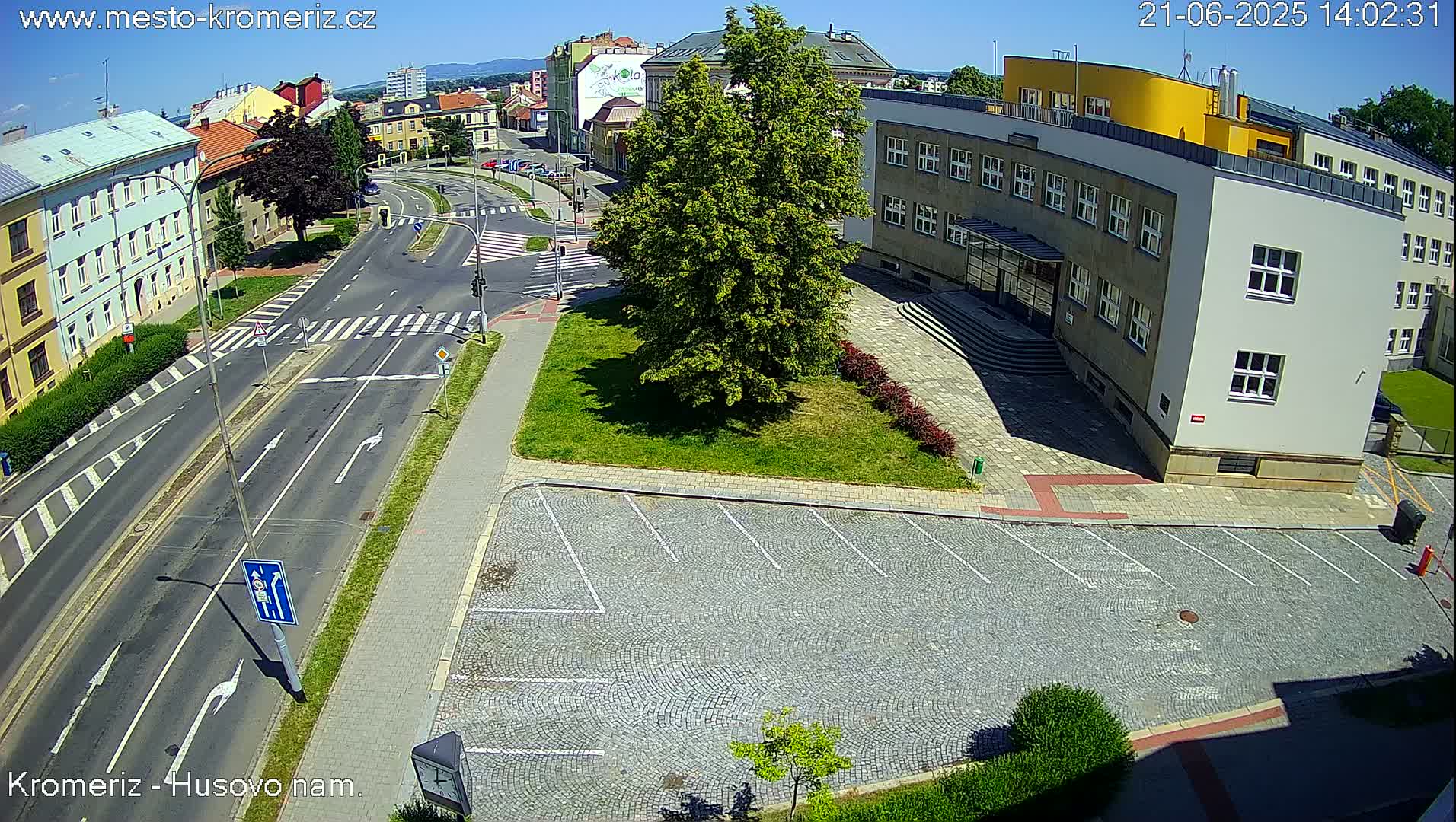 An aerial view of a street intersection on a sunny day, showing buildings, a grassy area with a tree, and a cobblestone parking lot.