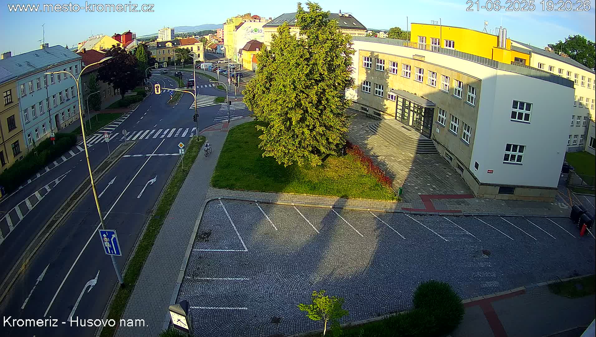 An aerial view of a street intersection with a cobblestone parking lot and buildings, under a mostly clear evening sky.