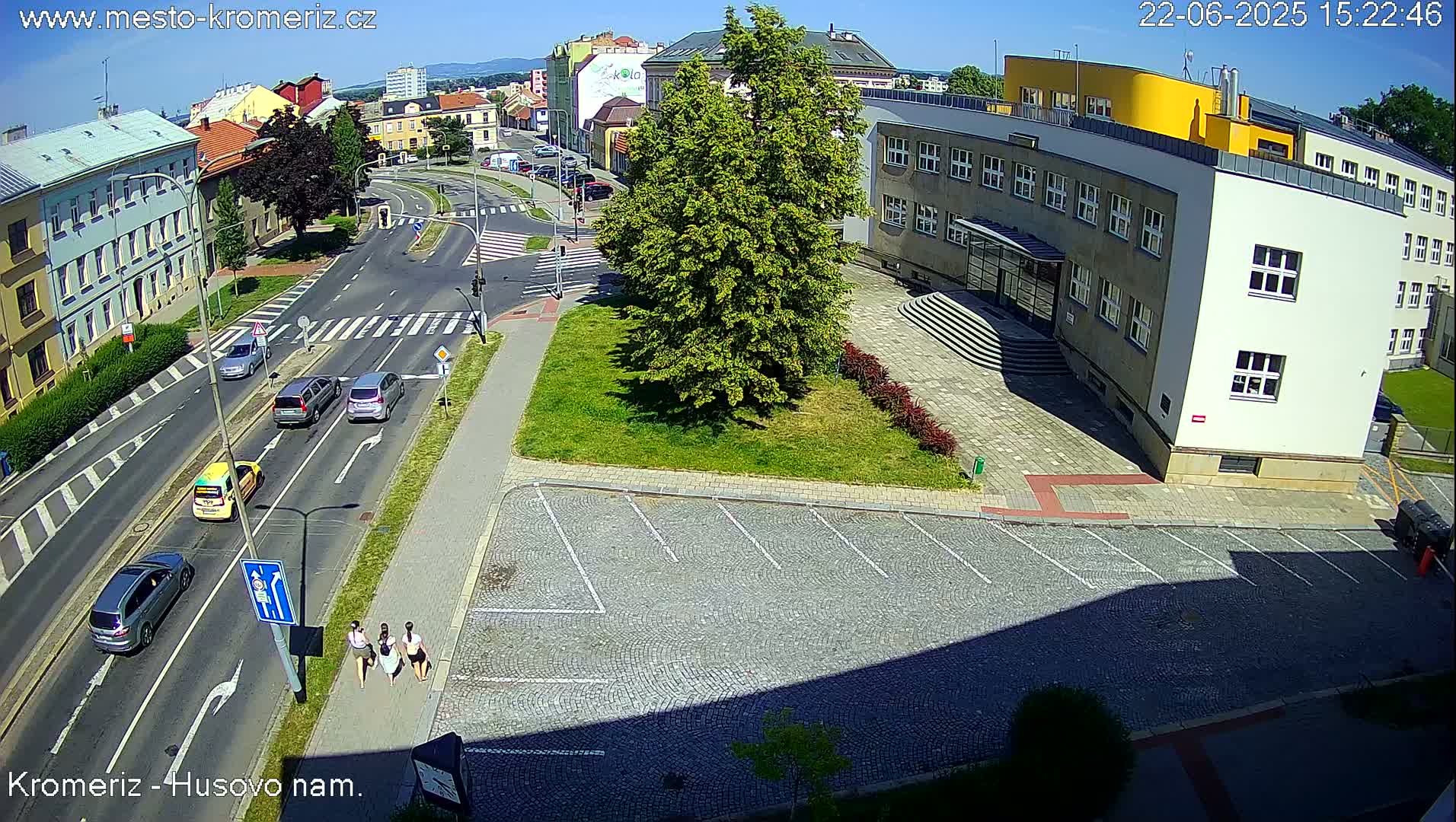 A sunny day view of a city street intersection with several cars, a group of pedestrians, and a large building with a cobblestone parking lot in front.