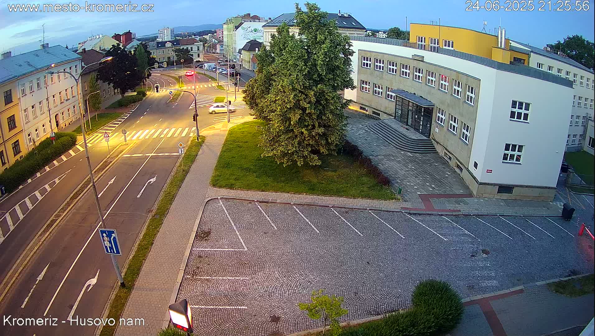 An empty cobblestone parking lot is adjacent to a street intersection at dusk, with a few cars visible on the road and buildings in the background.