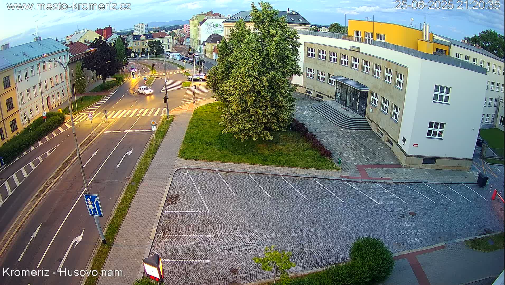 An empty cobblestone parking lot is adjacent to a street intersection and a multi-story building under a partly cloudy evening sky.