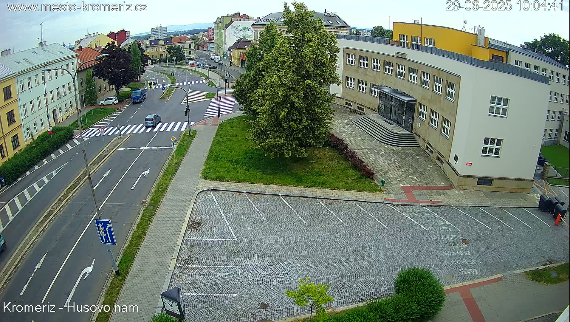 A street scene on an overcast day shows a two-lane road with crosswalks, buildings, a large tree, and an empty cobblestone parking lot.