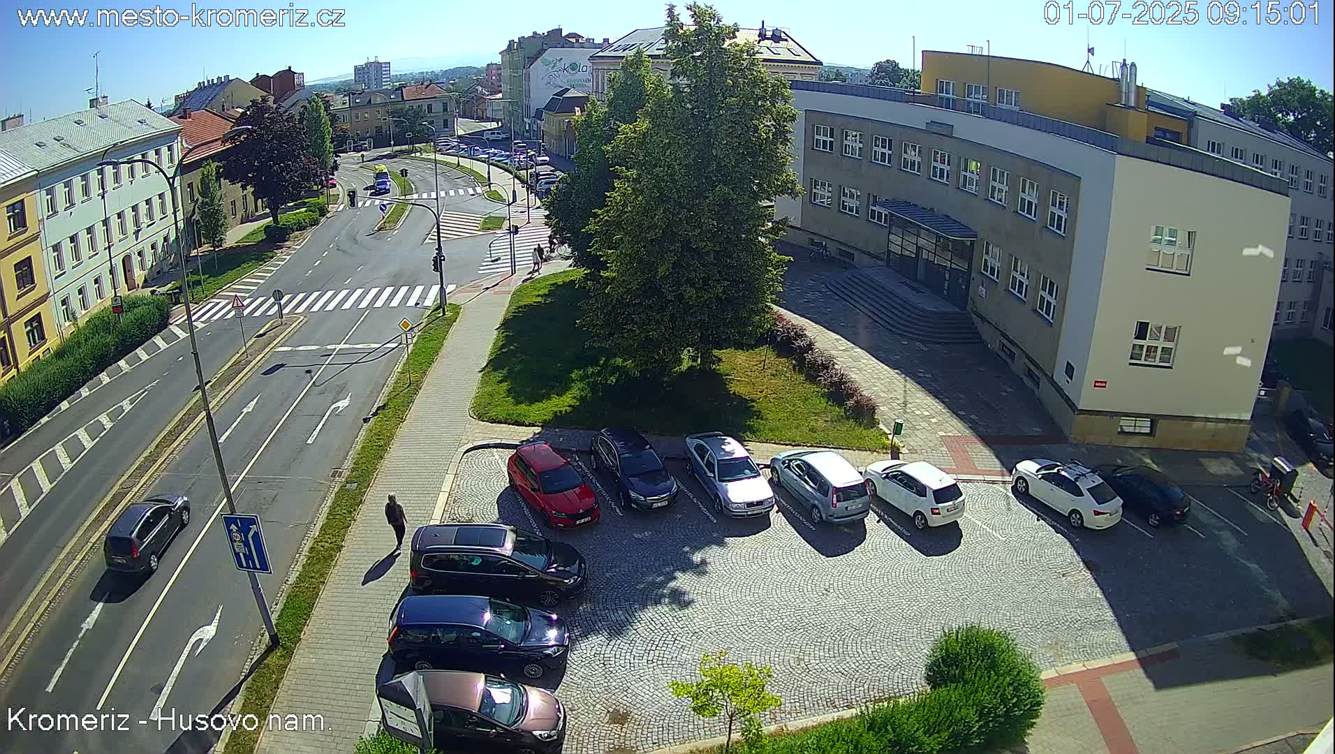 A sunny day view of a city intersection with several cars parked in a lot next to a large building, and other cars driving on roads.