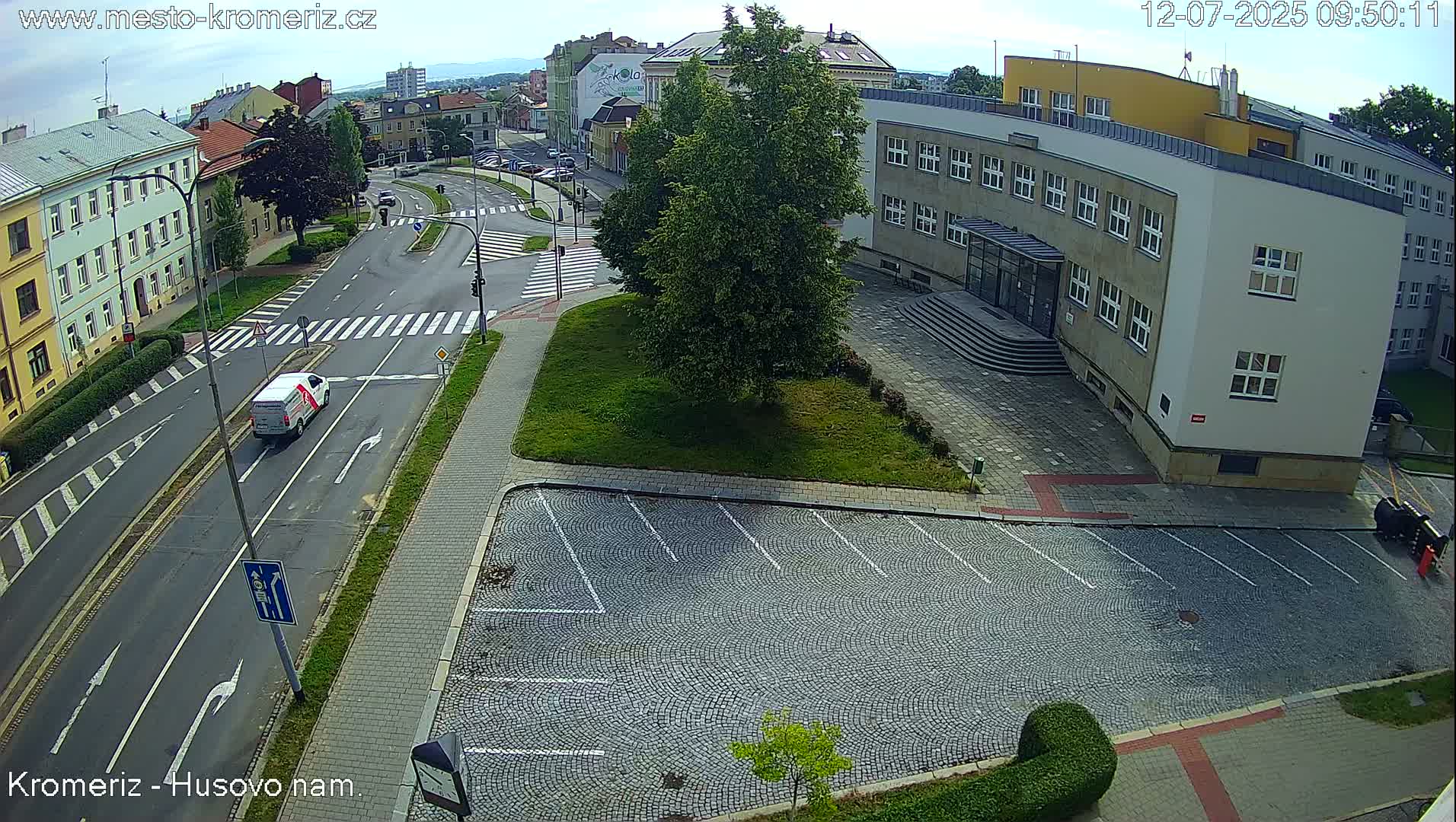 A mostly empty cobblestone parking lot sits beside a multi-story building next to a roadway with a white van driving on a sunny day.