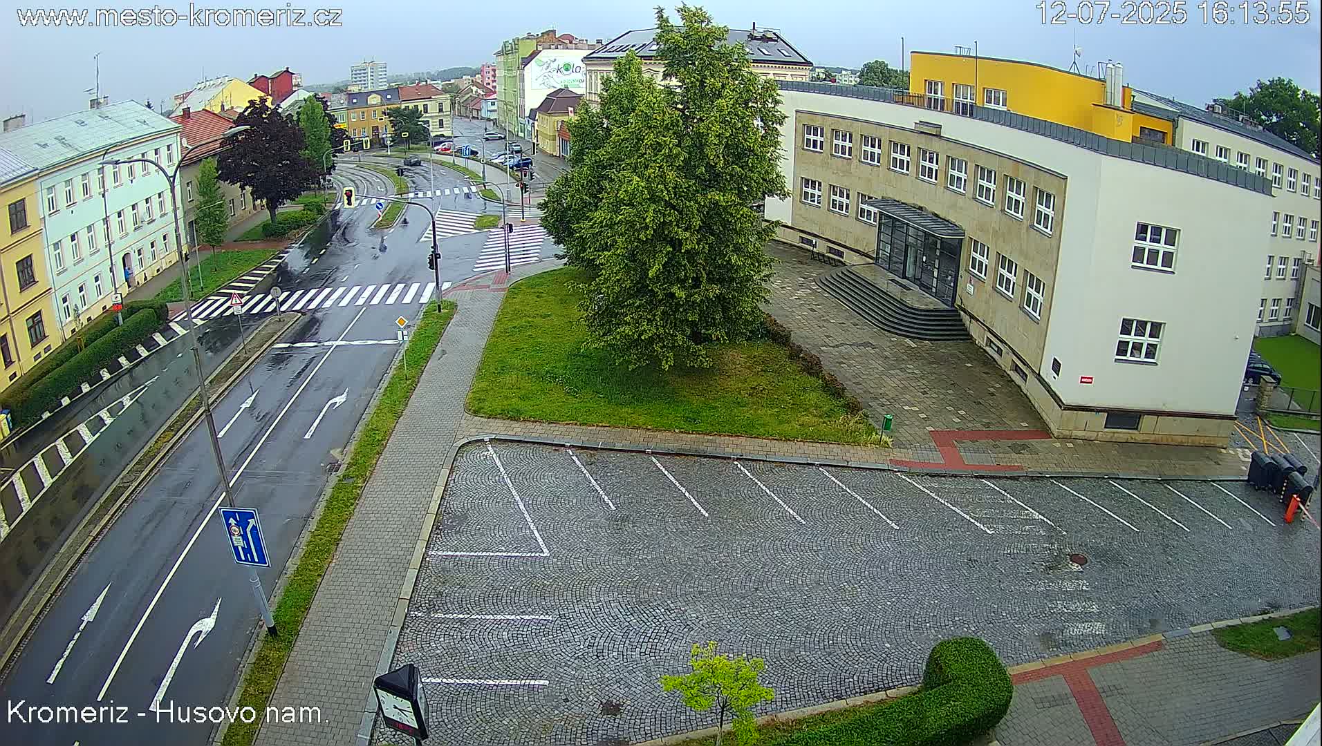 A mostly empty cobblestone parking lot sits beside a multi-story building next to a roadway with a white van driving on a sunny day.