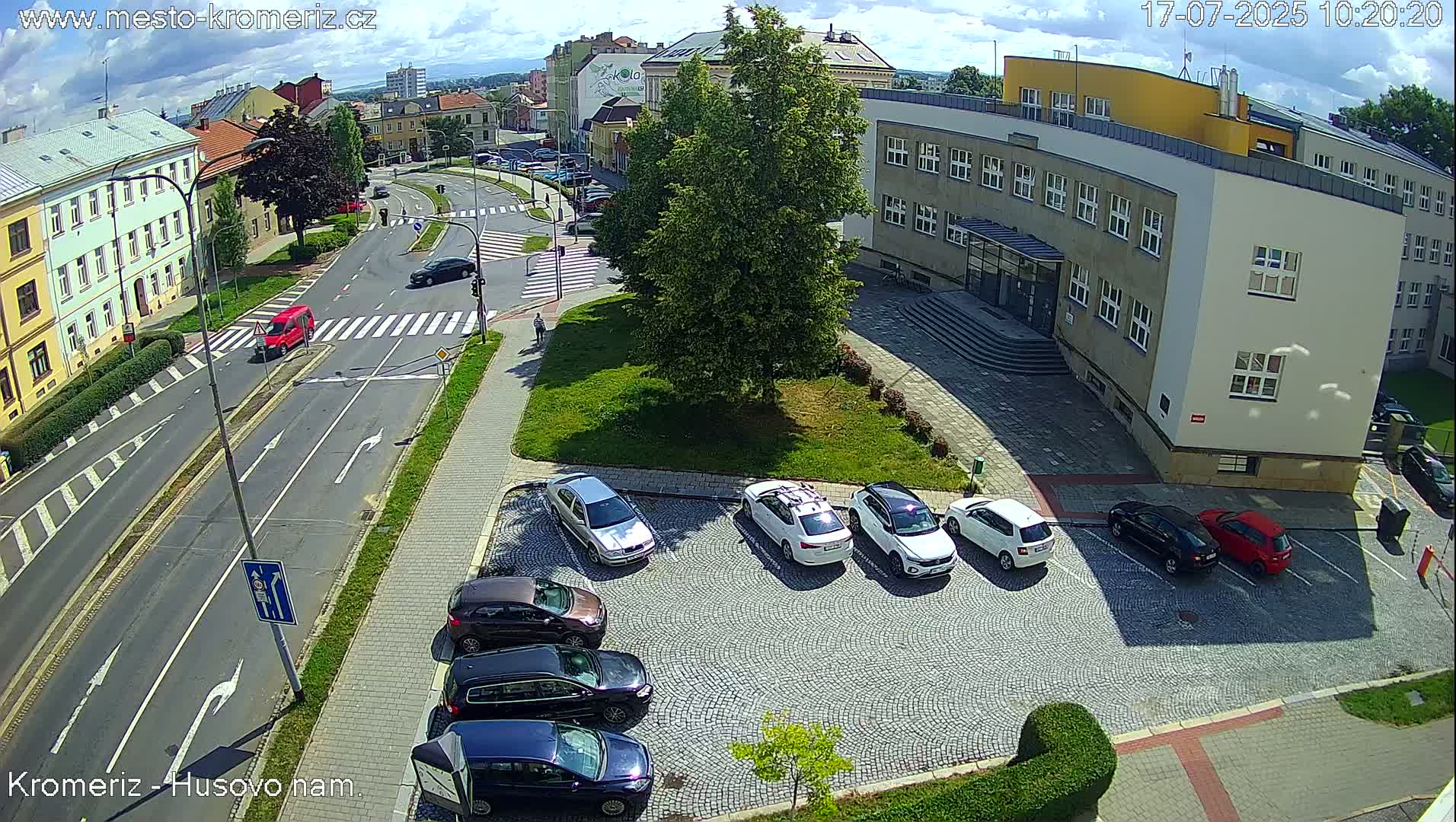 A sunny day reveals a street scene with several cars parked in a lot next to a large building, and other vehicles driving on the roads near a pedestrian crossing.