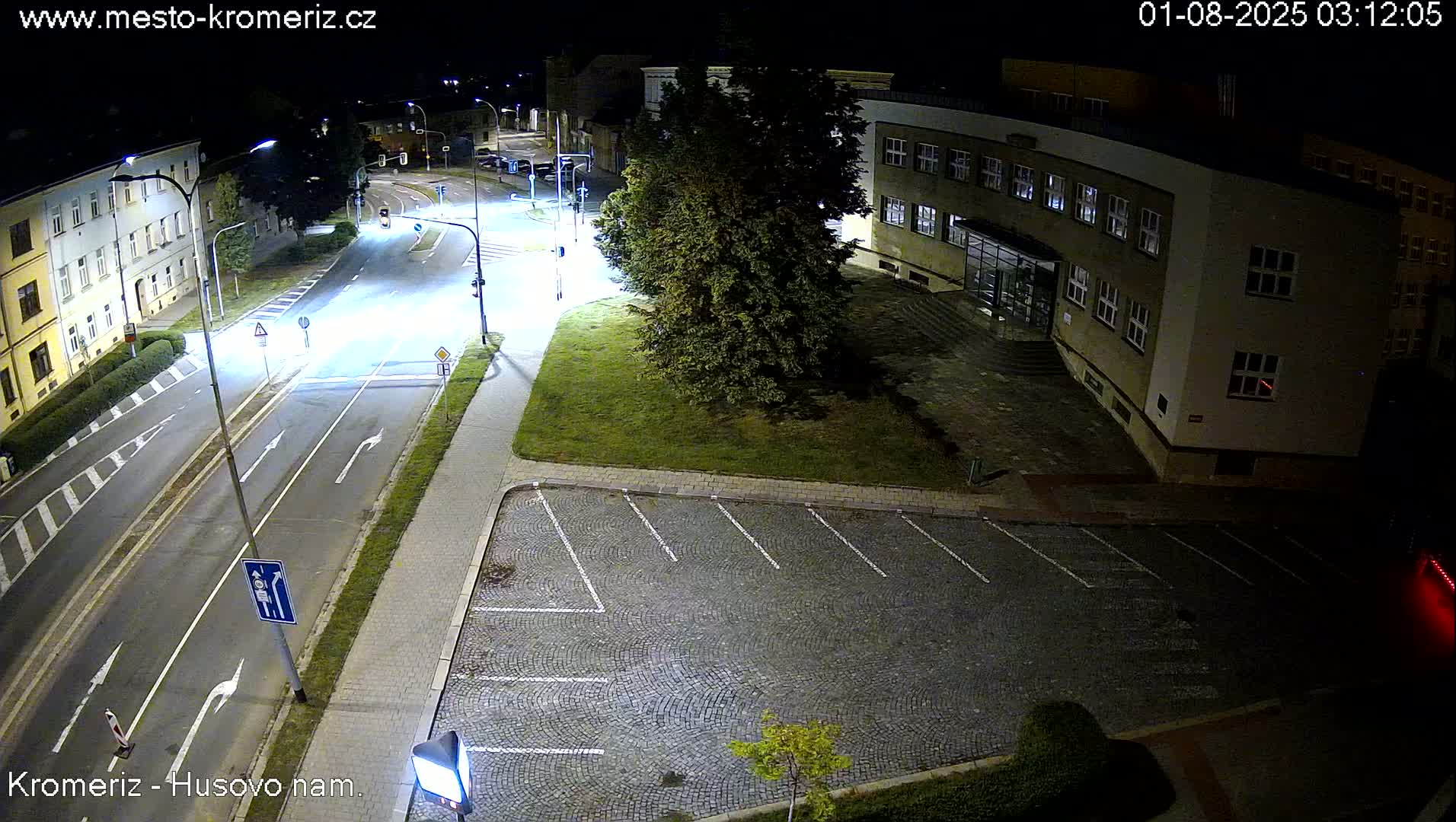 A nighttime view of a paved street and empty parking lot next to a multi-story building, illuminated by streetlights.