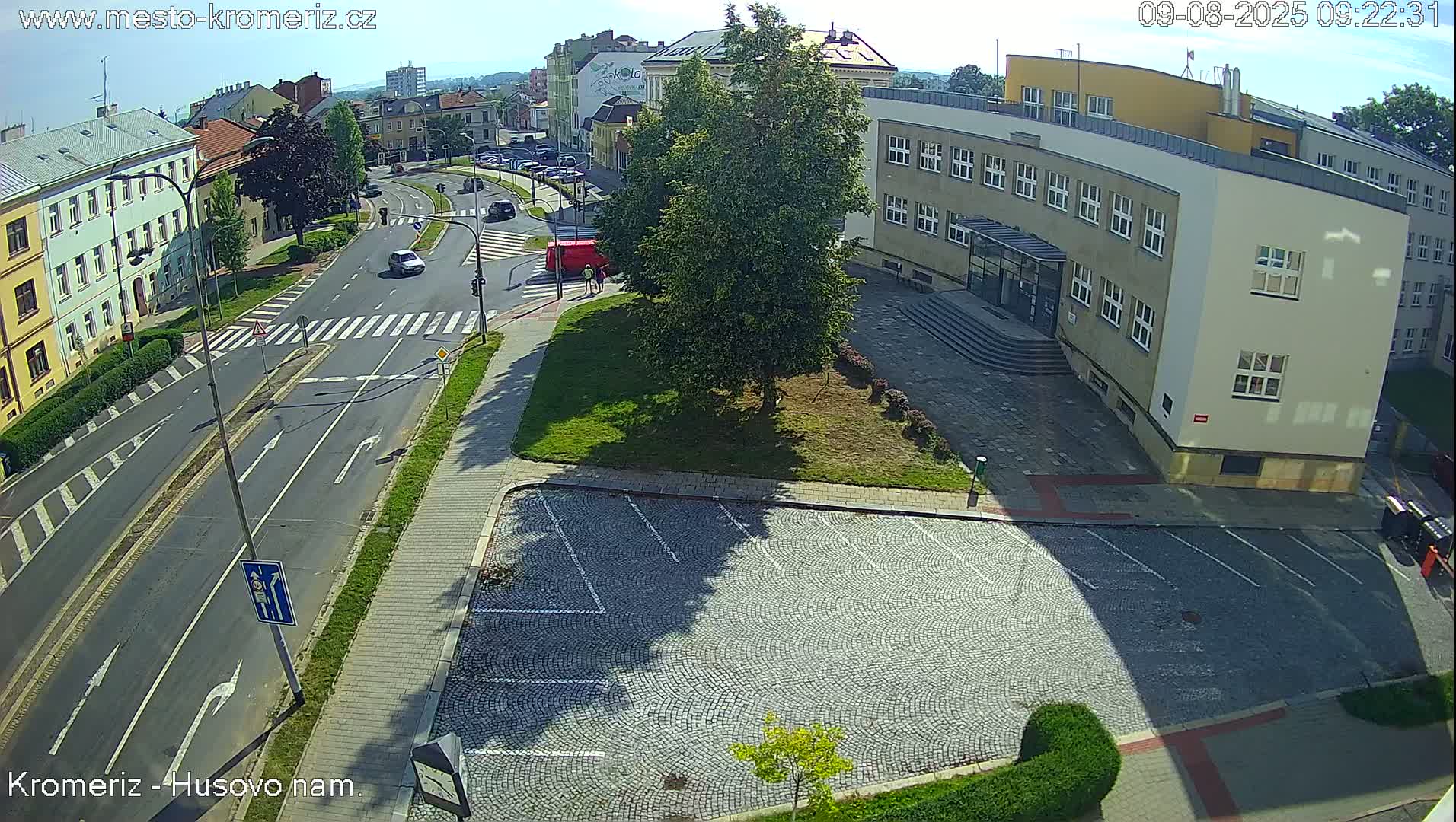 A sunny view of a paved street intersection with several buildings, a small grassy area, and an empty cobblestone parking lot.