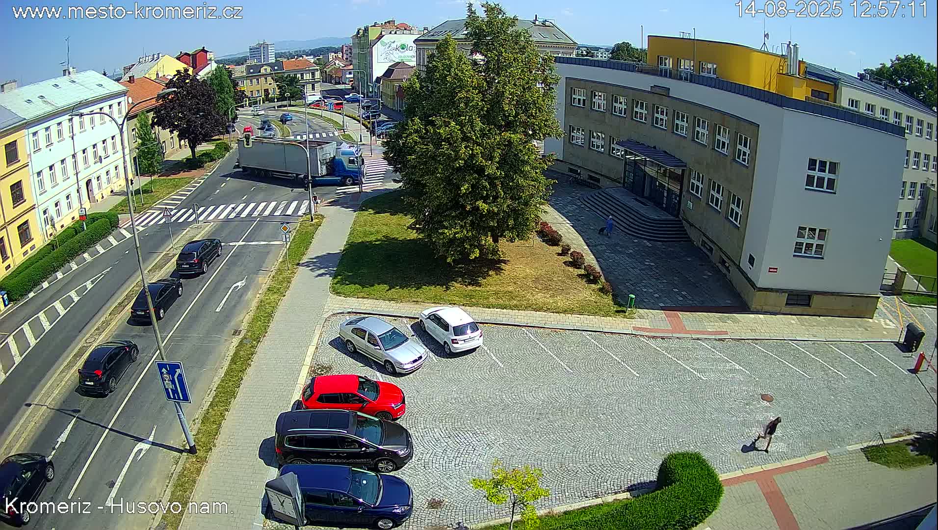 A sunny view of a paved street intersection with several buildings, a small grassy area, and an empty cobblestone parking lot.