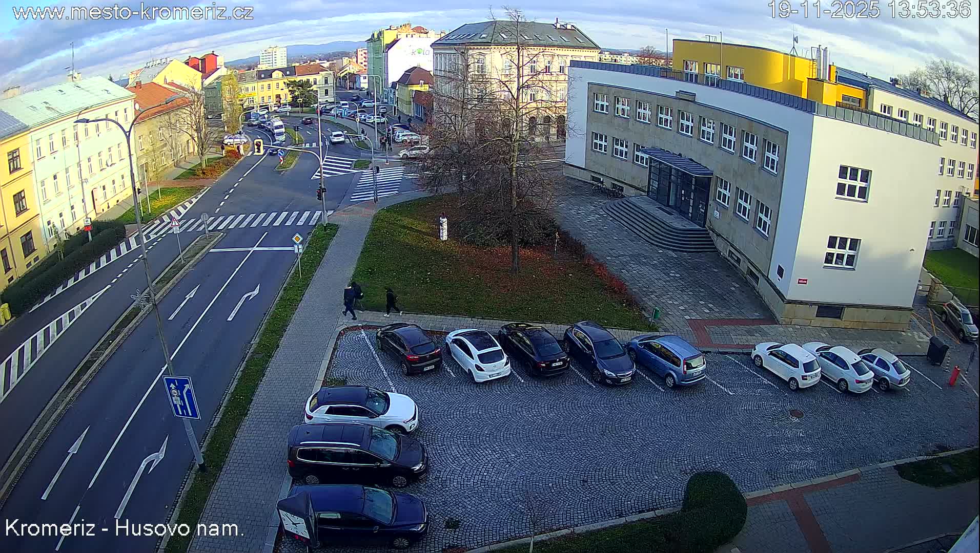 A sunny view of a paved street intersection with several buildings, a small grassy area, and an empty cobblestone parking lot.