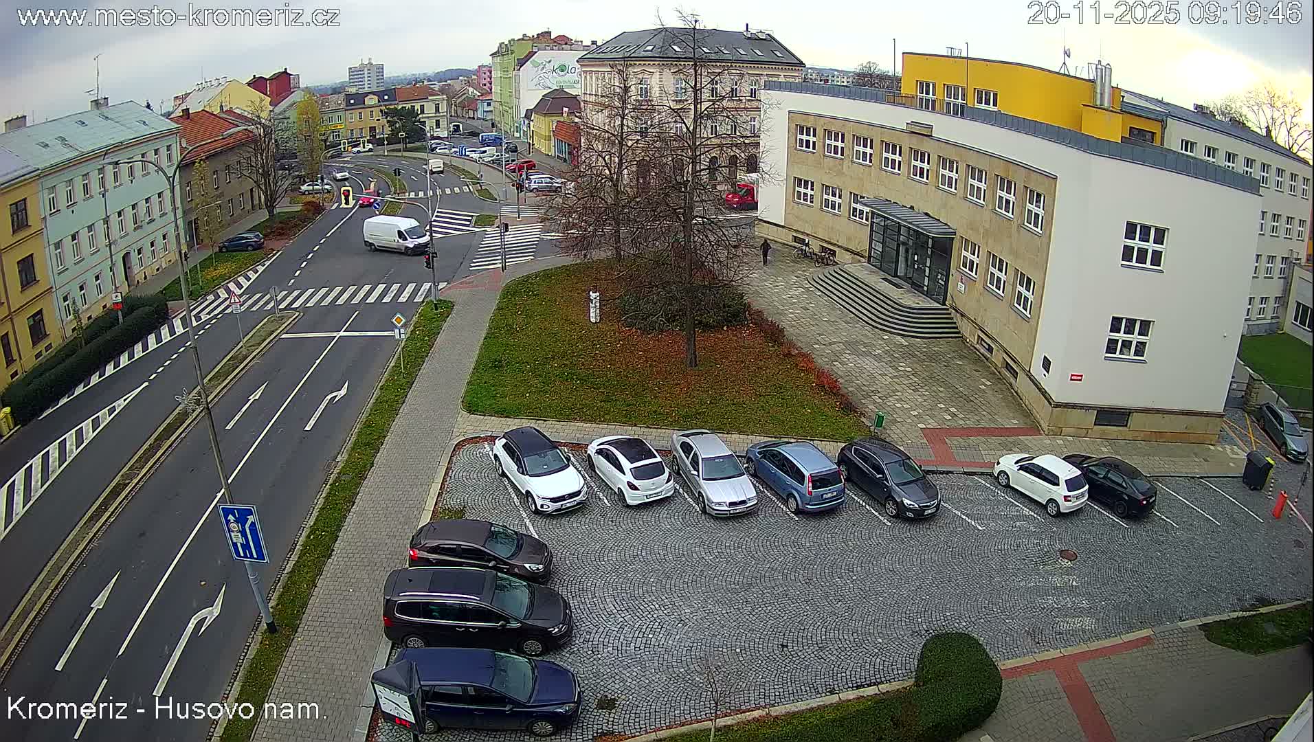 An overhead view reveals an urban street scene featuring a busy intersection with cars and a white van, multiple buildings including one with a distinctive yellow upper section, a cobblestone parking lot filled with vehicles, and bare trees in a green space, all beneath an overcast sky.