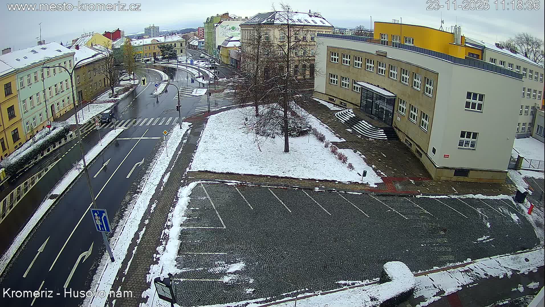 An overcast winter day reveals a quiet European town scene with wet roads and sidewalks, patches of snow covering the ground and rooftops, and bare trees surrounded by various multi-story buildings.