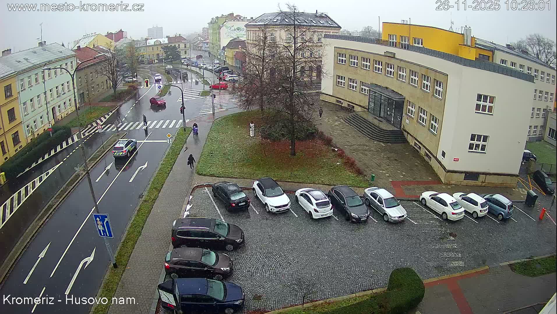 An elevated view reveals a wet city street with cars and pedestrians, bordered by buildings and a cobblestone parking lot, beneath an overcast sky.
