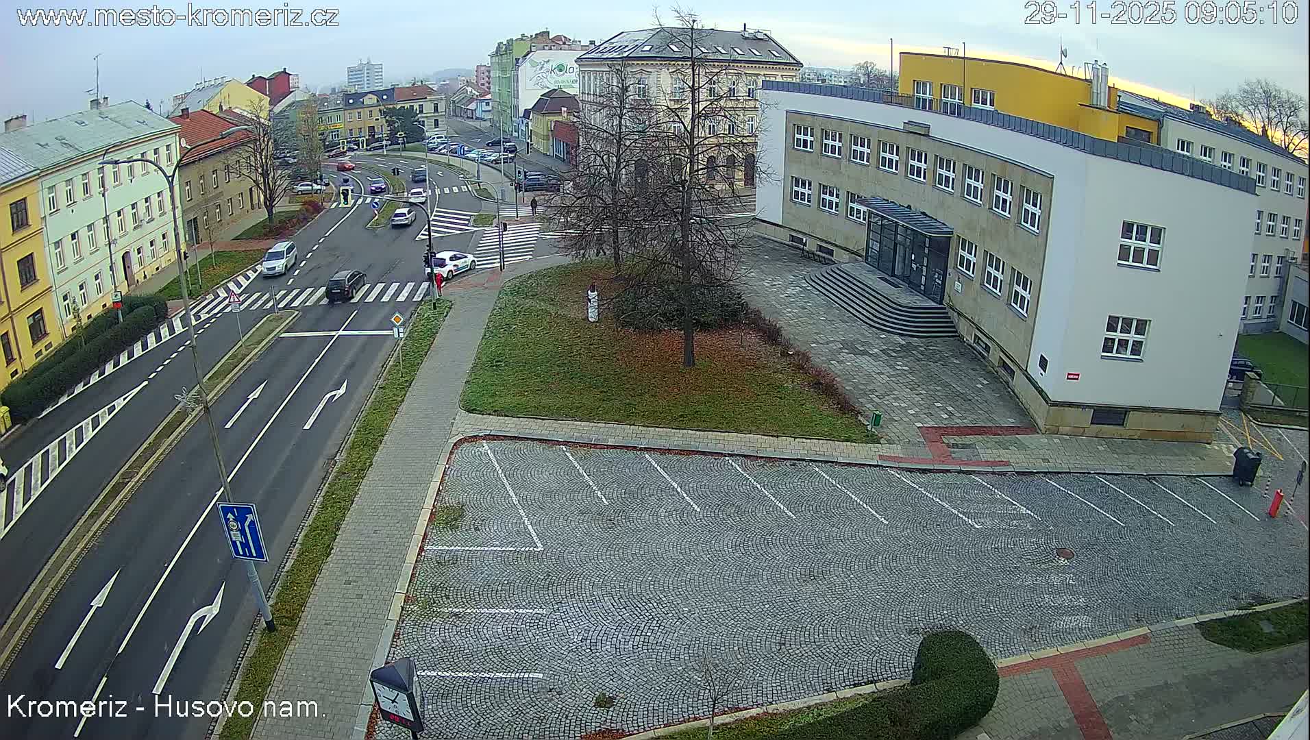 An overcast day depicts an urban street intersection with several cars and a few pedestrians, flanked by diverse buildings including a large modern structure, and a largely empty cobblestone parking lot.